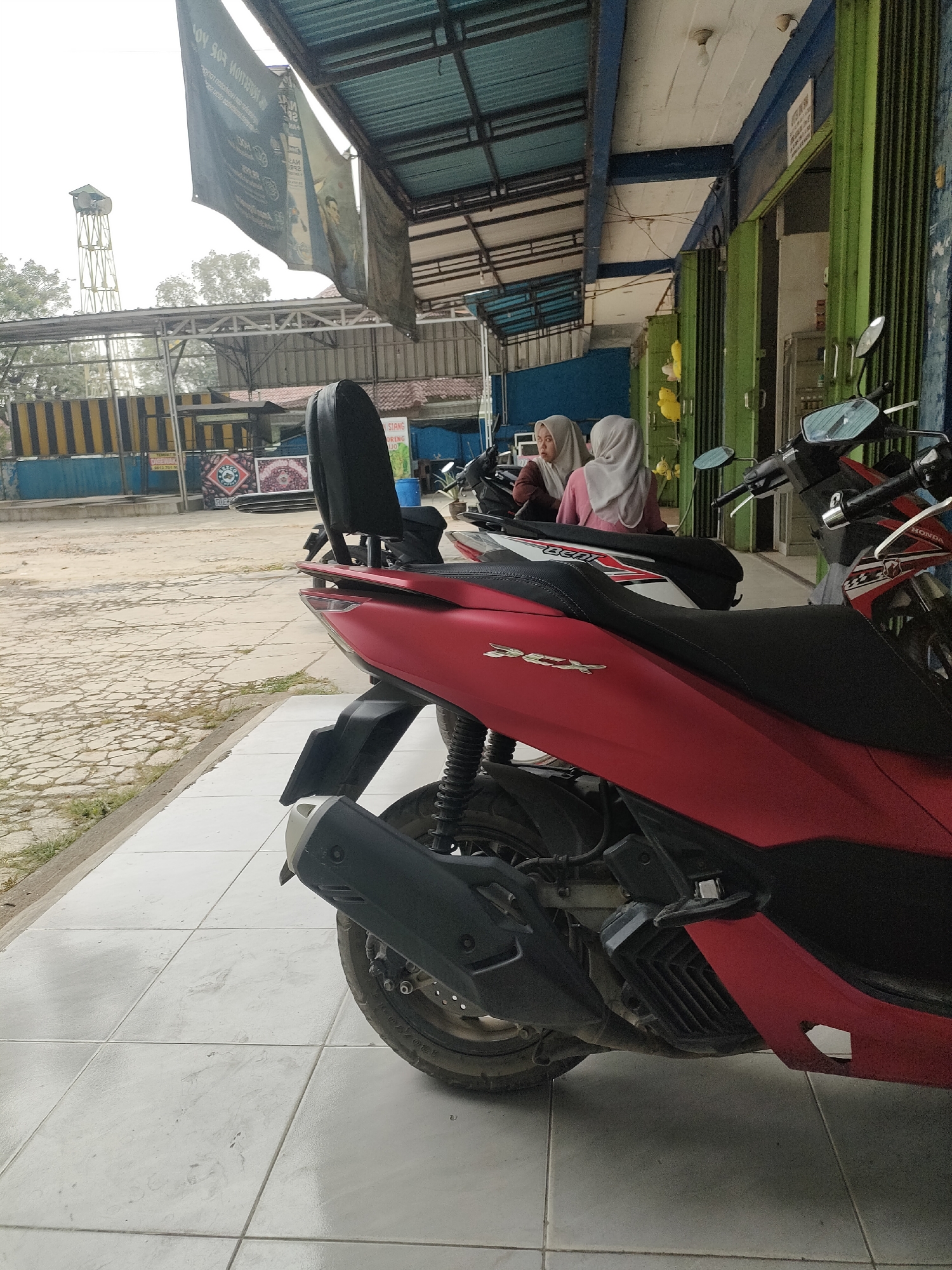 A red and black scooter is parked in the foreground on a tiled walkway. The scooter has a black seat, a black backrest, and "PCX" is embossed on its red body. To the left of the scooter, a white and black scooter is partially visible. Two women wearing hijabs are seated on the white and black scooter. One woman wears a light gray hijab and a pink shirt, and the other wears a dark gray hijab. In the background, a building with green doors and windows is visible. A banner hangs from a metal structure above the building. Beyond the building, a water tower and trees are seen under an overcast sky. The walkway is made of large light-colored tiles, and a rougher, cracked pavement is visible behind it. The scene appears to be outdoors, possibly in a commercial or public area in Pringsewu, Indonesia.