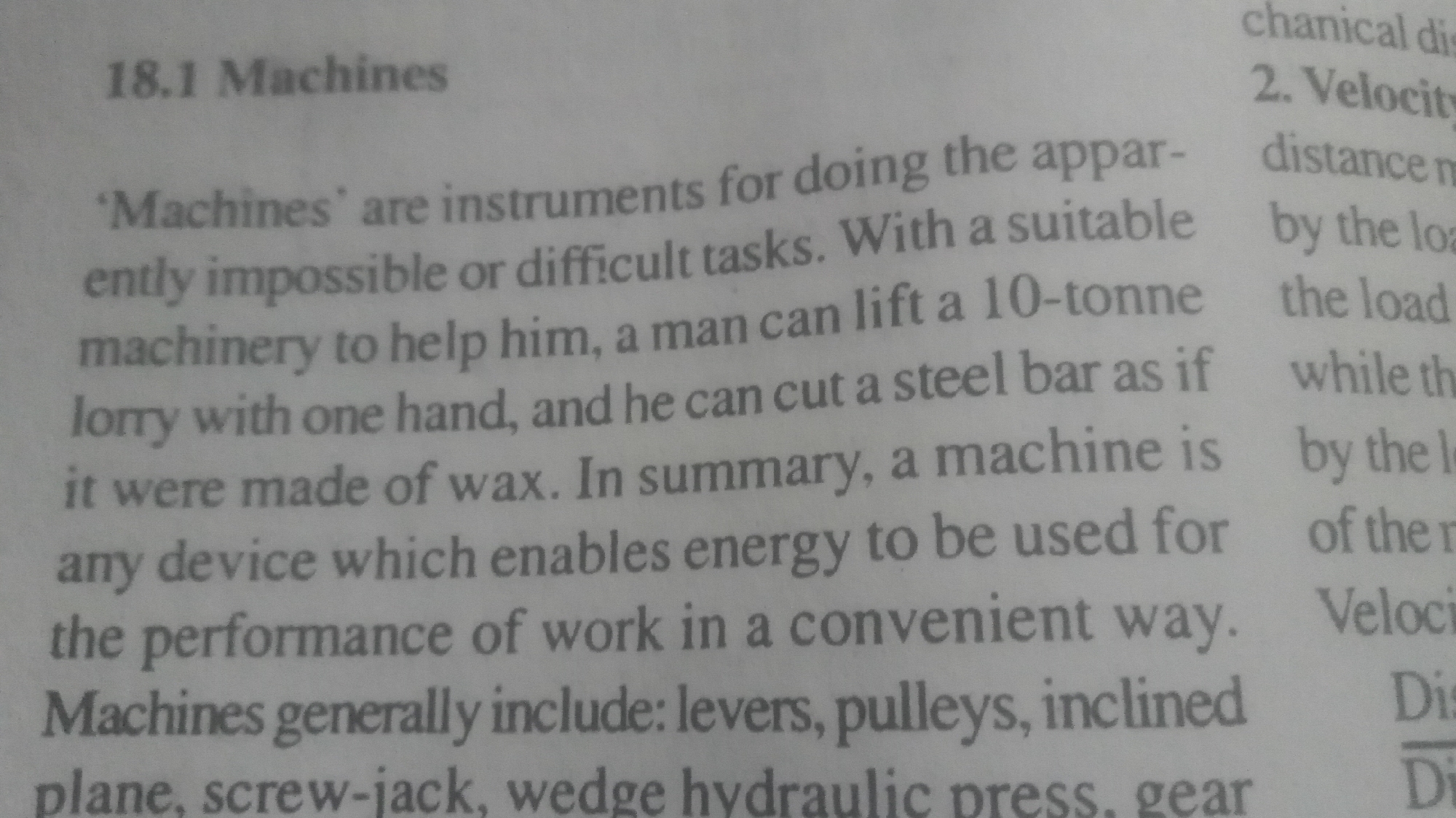 The image is a close-up photograph of a textbook page titled "18.1 Machines." The text defines machines as instruments for doing apparently impossible or difficult tasks. It uses the example of a man lifting a 10-tonne lorry with one hand or cutting a steel bar as if it were wax with the help of suitable machinery. A machine is summarized as any device that enables energy to be used for work in a convenient way. The text mentions that machines generally include levers, pulleys, inclined planes, screw-jacks, wedge hydraulic presses, and gears. The background is a plain white surface, which is the textbook page.
On the right side of the image, partially obscured, there are words about mechanical distance and velocity. The image is indoors and well-lit, suggesting it was taken in a classroom or study setting.