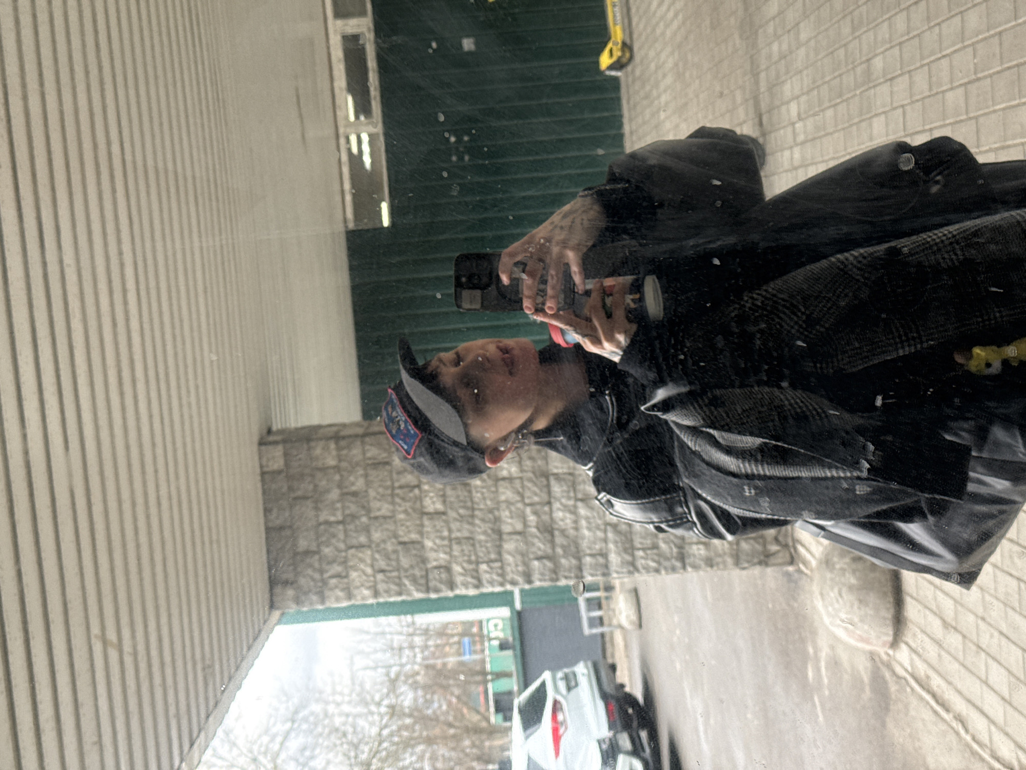 A person is taking a selfie in a reflection. The person is wearing a black cap with an emblem, a black shirt, and a black leather jacket over a plaid jacket. They have tattoos on their hands. They are holding a smartphone. The reflection shows a street with a white van, a building with white siding, and a brick facade. The image was taken in Saint Petersburg, Russia.
