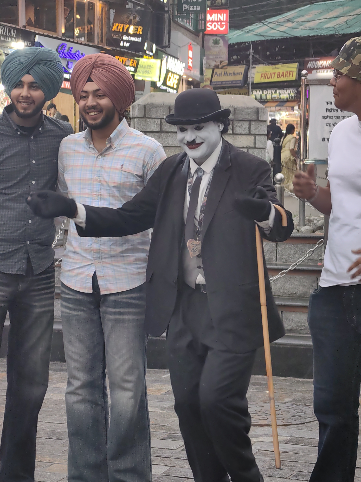 The image depicts a street scene in Manali, India, likely near the Mall Road, a popular tourist area known for its shops and eateries. In the foreground, a person in Charlie Chaplin-esque makeup and attire, complete with a bowler hat, white face paint, dark suit, and a cane, stands center frame. To the left, two young men wearing turbans, one in a dark patterned shirt and the other in a plaid shirt, are positioned close to the Chaplin impersonator. The man in the plaid shirt is smiling. To the right, another individual in a white t-shirt and a camouflage-patterned hat is visible, seemingly interacting with the Chaplin impersonator. The background shows various signs for businesses, including "KHYBER Family Restaurant & Bar," "Ballantine's," "Madras Cafe," and "Fruit Barfi," suggesting a commercial and bustling environment. The ground appears to be paved with stone. The lighting suggests it is daytime.