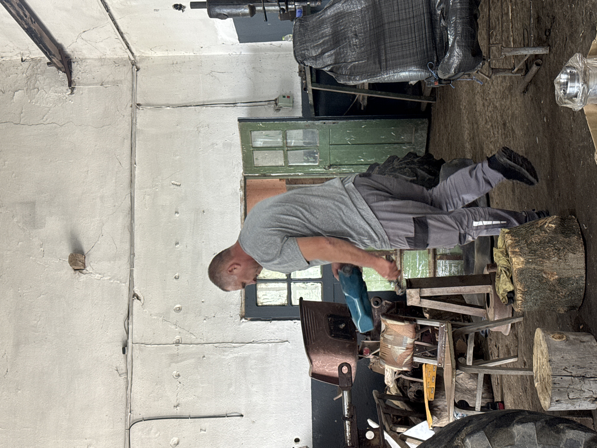 The image captures a man working in a rustic workshop setting, likely in Sarnevo, Bulgaria. The man, appearing to be middle-aged with short hair, wears a grey t-shirt and grey work pants with black accents. He is actively engaged, leaning forward and holding a blue power tool, possibly a drill or grinder, focused on a task at a wooden workbench or piece of equipment in front of him.
The workshop has bare, off-white concrete or plastered walls exhibiting visible cracks and wear, along with exposed electrical conduits and wiring. A dark green wooden door with four glass panes is prominent in the background. The floor is an earthy or concrete surface, cluttered with various tools and materials. Several tree stumps serve as makeshift seats or stands, and a simple wooden chair with a patterned cushion is also present. Other visible objects include a white electrical control box on the wall with red and green buttons, a black tarp covering some machinery, and a metallic cylindrical part wrapped in clear plastic. A large black tire is partially visible in the foreground. The scene suggests a daytime setting due to the overall brightness, and the environment is typical of a functional, unrefined workspace, possibly for mechanical repairs or crafting. No discernible text is visible.
