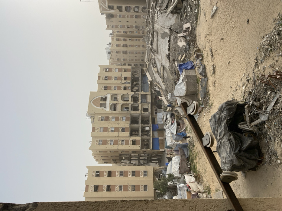 The image depicts a severely damaged urban area in Khan Yunis, Palestinian Territory, viewed from an elevated position. In the background, multiple multi-story buildings, likely apartment blocks of light beige color, exhibit extensive structural damage, including numerous impact marks on facades and blown-out windows. The lower floors of the central buildings are largely destroyed, revealing interior wreckage. The ground in the midground and foreground is covered with significant piles of concrete rubble, twisted metal, and sand. Amidst this debris and open sandy patches, a cluster of approximately a dozen makeshift shelters, constructed from tarpaulins (white, grey, blue, and brown) stretched over frames, are visible. Two individuals are present among these temporary structures; one person in dark clothing is walking left-to-right near a dark tarp, and another lighter-colored figure is further back. In the immediate foreground, a rusted metal beam extends horizontally from the left, fitted with three white pulleys. Below this beam, a pile of rubble and a dark tarp-covered object are present. The sky is overcast and hazy.