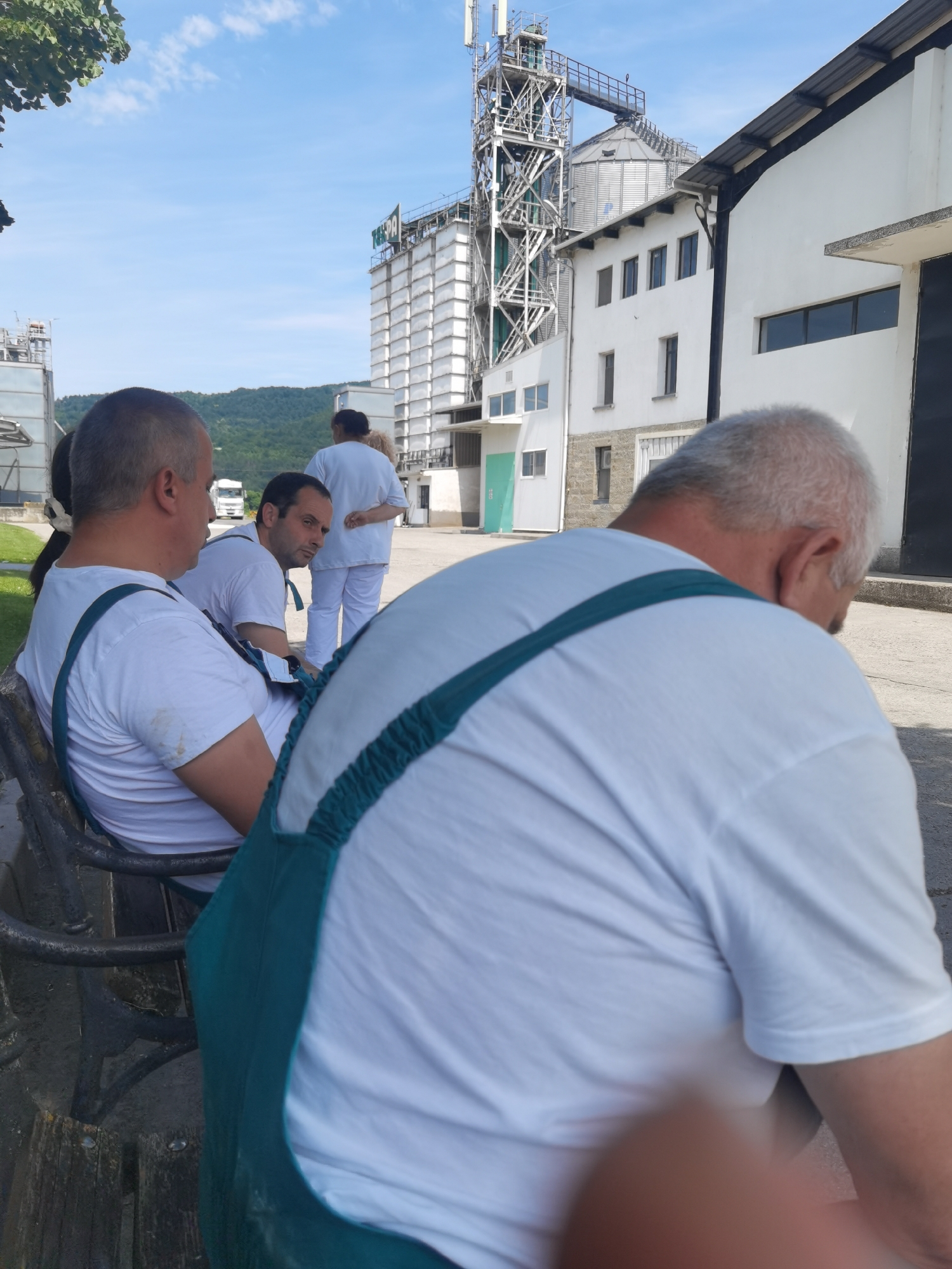 The image depicts an outdoor scene at an industrial facility, likely a grain silo complex, in Lomets, Bulgaria. In the foreground, three individuals are present, two of whom are seated on a dark-colored bench. They are wearing white t-shirts and green suspenders. The person on the far right is partially visible, with their back to the camera, showing a white t-shirt and green suspenders. To their left, another individual is seated, looking towards the right, also in a white t-shirt and green suspenders. To the left of these two, a third individual is seated, looking forward, wearing a white t-shirt and green suspenders, with a dark object on the bench beside them.
In the background, a white building with several windows is visible, adjacent to a tall, metallic industrial structure that resembles a grain elevator with conveyor belts and platforms. Further in the background, a truck is parked on a paved area. Another person in white attire is walking away from the viewer towards the building. The sky is blue with scattered white clouds. The ground appears to be a paved surface. The overall impression is a working environment, possibly during a break, given the presence of people seated on a bench.