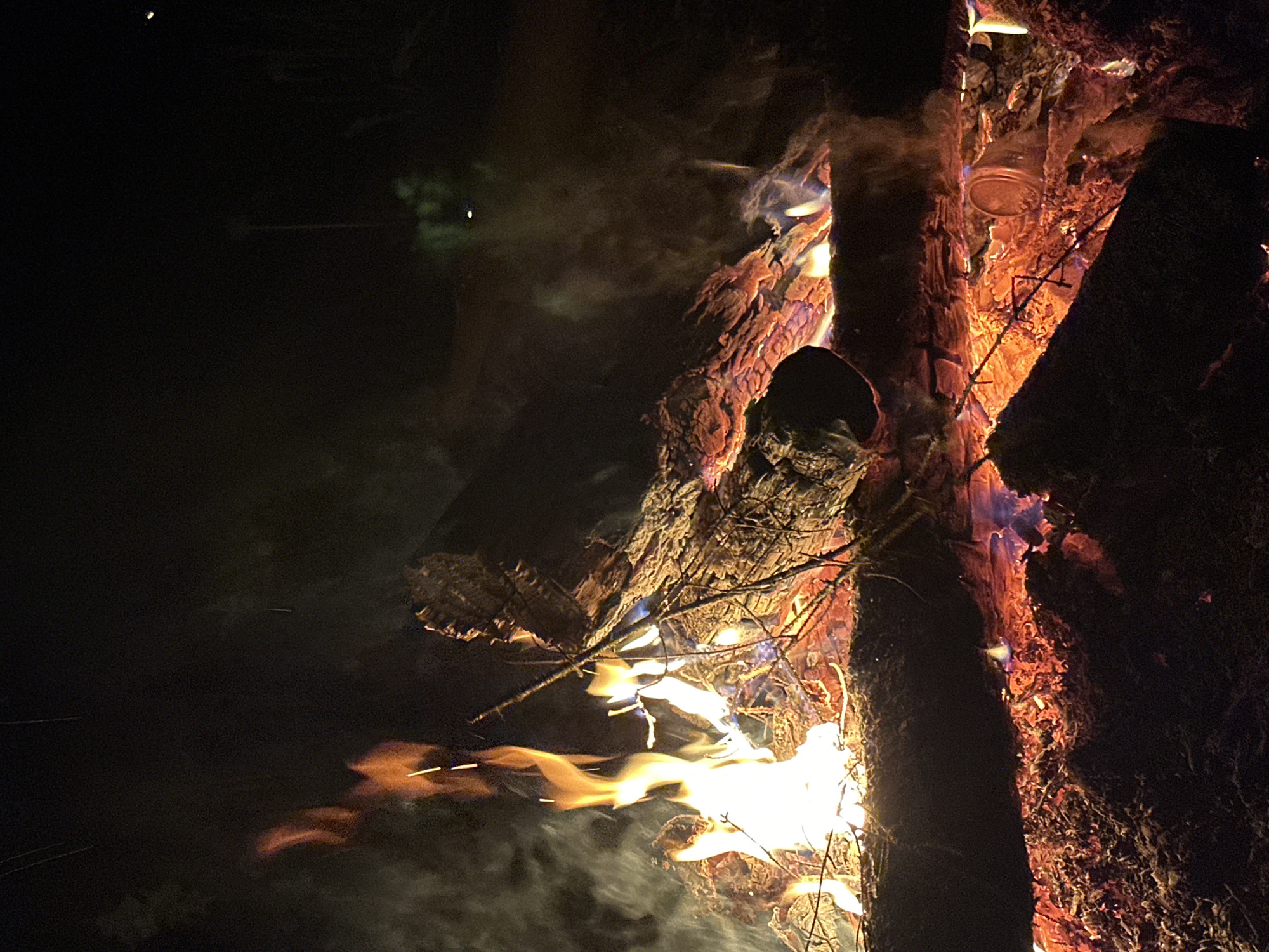 A bonfire burns with a medium-intensity fire. The location is not discernible from the image. The bonfire is composed of logs and branches, with flames and smoke emanating from within. There are no people present in the image. A jar is resting upon the wood and metal implements are used to help the jar stand. The surrounding environment appears to be dark.