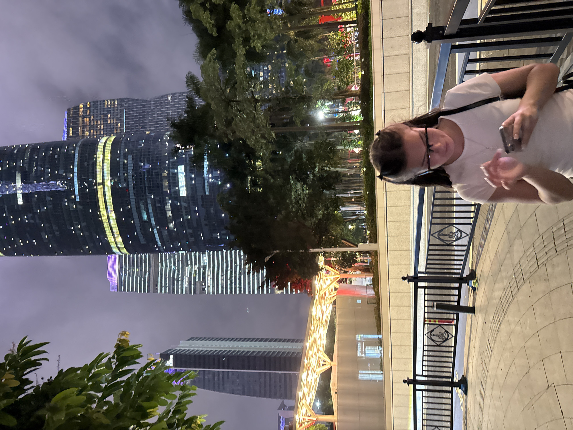 A young woman with dark hair pulled into two pigtails, wearing glasses and a white t-shirt, is looking down at her mobile phone, which she is holding in her hands. She appears to be walking or standing on a paved walkway in an urban setting at night. Behind her, the illuminated modern skyscrapers of Tianhe District, Guangzhou, China, are visible, some with purple and yellow lighting. The sky is overcast. A black metal fence runs along the walkway in front of the woman. Trees and lush foliage are interspersed between the buildings and along the walkway. A covered walkway with orange lighting is visible in the middle ground.