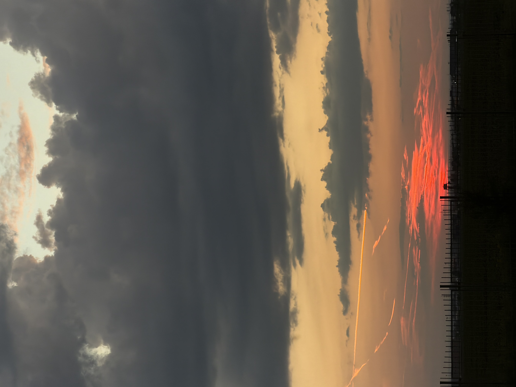 The image depicts a sunset over a rural landscape, possibly in or near the village of Timofeyevka, Russia. The sky is filled with dramatic clouds, transitioning from dark gray at the top to vibrant orange and red hues near the horizon. Multiple contrails are visible, streaking across the sky, some illuminated by the setting sun. In the foreground, a dark silhouette of a metal fence runs horizontally across the bottom of the frame, with a field of dark vegetation behind it. Beyond the field, a faint outline of industrial or agricultural structures can be seen against the brightly colored sky. The overall scene conveys a sense of atmospheric drama and the fading light of day. There are no people or discernible animals present.