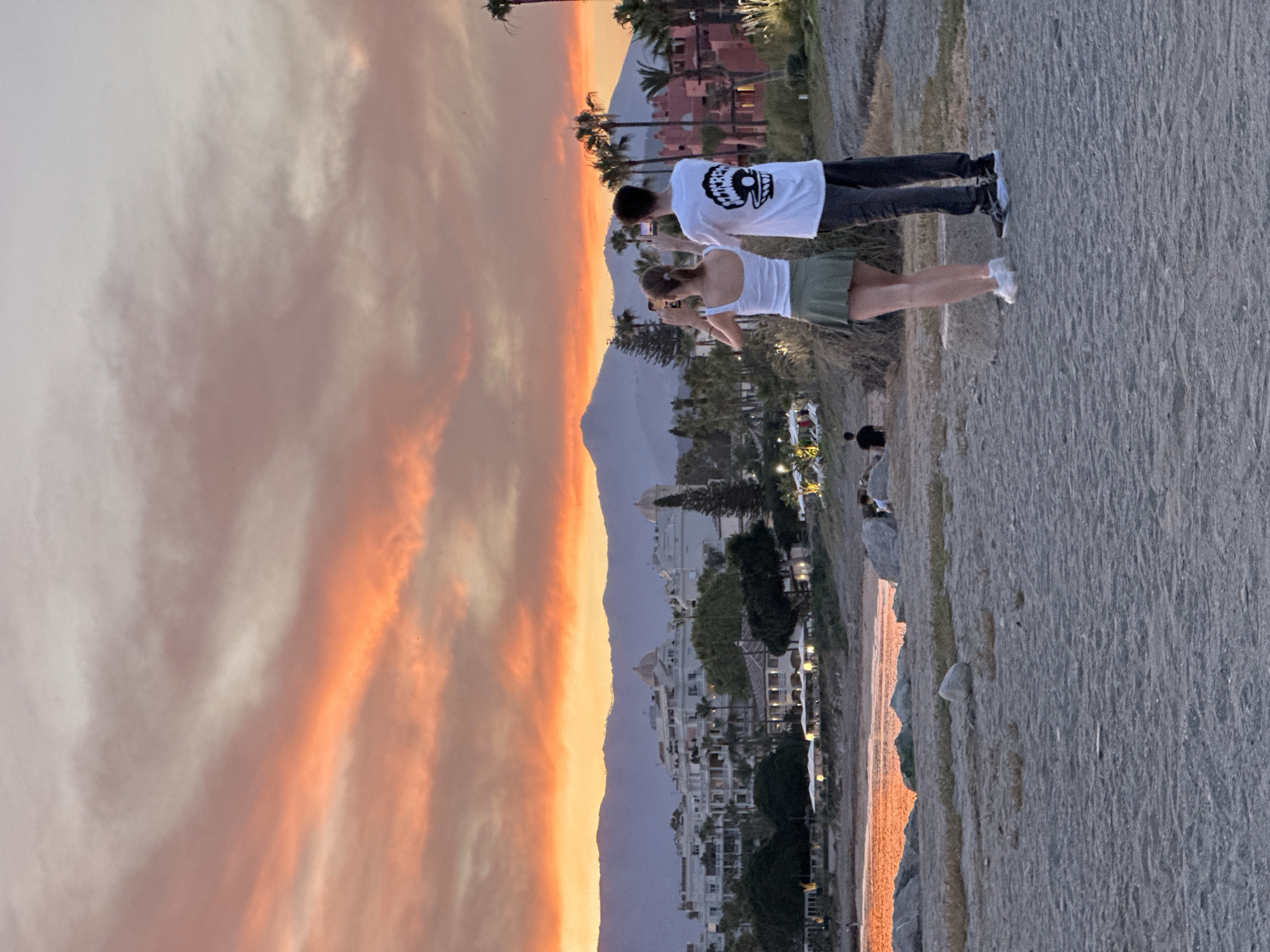Two people are standing on a beach in El Velerín, Spain, taking pictures of a sunset over the Mediterranean Sea. The sky is filled with vibrant orange and pink clouds. In the background, there is a town with white buildings and palm trees. The sea reflects the colors of the sunset. The beach is sandy and there are rocks scattered on the shore. The people appear to be a couple, both looking out at the view and holding up their phones to capture the moment. The man is wearing a white t-shirt with a graphic print and dark pants, while the woman is wearing a white tank top and a green skirt. The overall scene is peaceful and romantic, capturing the beauty of a Spanish coastal sunset.