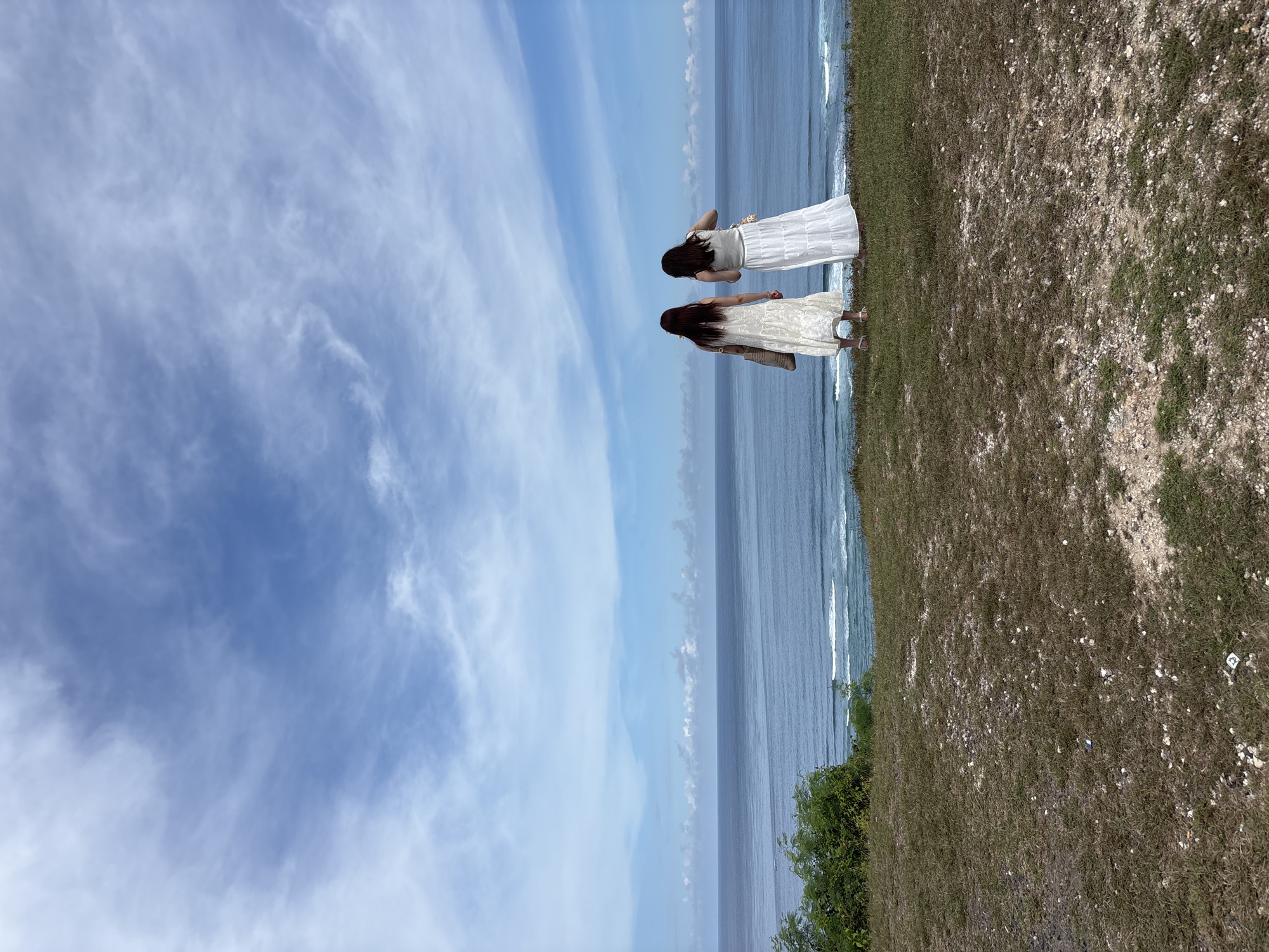 Two women stand on a grassy cliff overlooking the ocean under a blue sky with scattered clouds. The location is Kuta Selatan, Indonesia. Both women have long dark hair and are wearing light-colored dresses. One is looking out towards the ocean. Vegetation grows on the edge of the cliff.