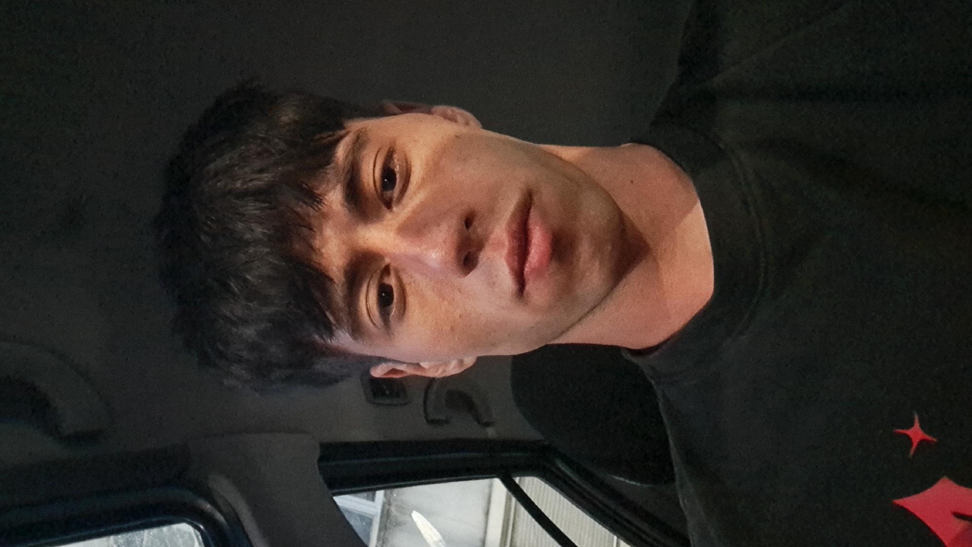 A close-up selfie of a young man with dark, tousled hair and dark eyes is taken inside a car. He is wearing a black t-shirt with a red graphic design on the lower left side. The man is looking directly at the camera with a neutral expression. The interior of the car is dimly lit, with a portion of the window visible on the left side of the image, showing an out-of-focus background that appears to be urban. The location is identified as Burgas, Bulgaria.