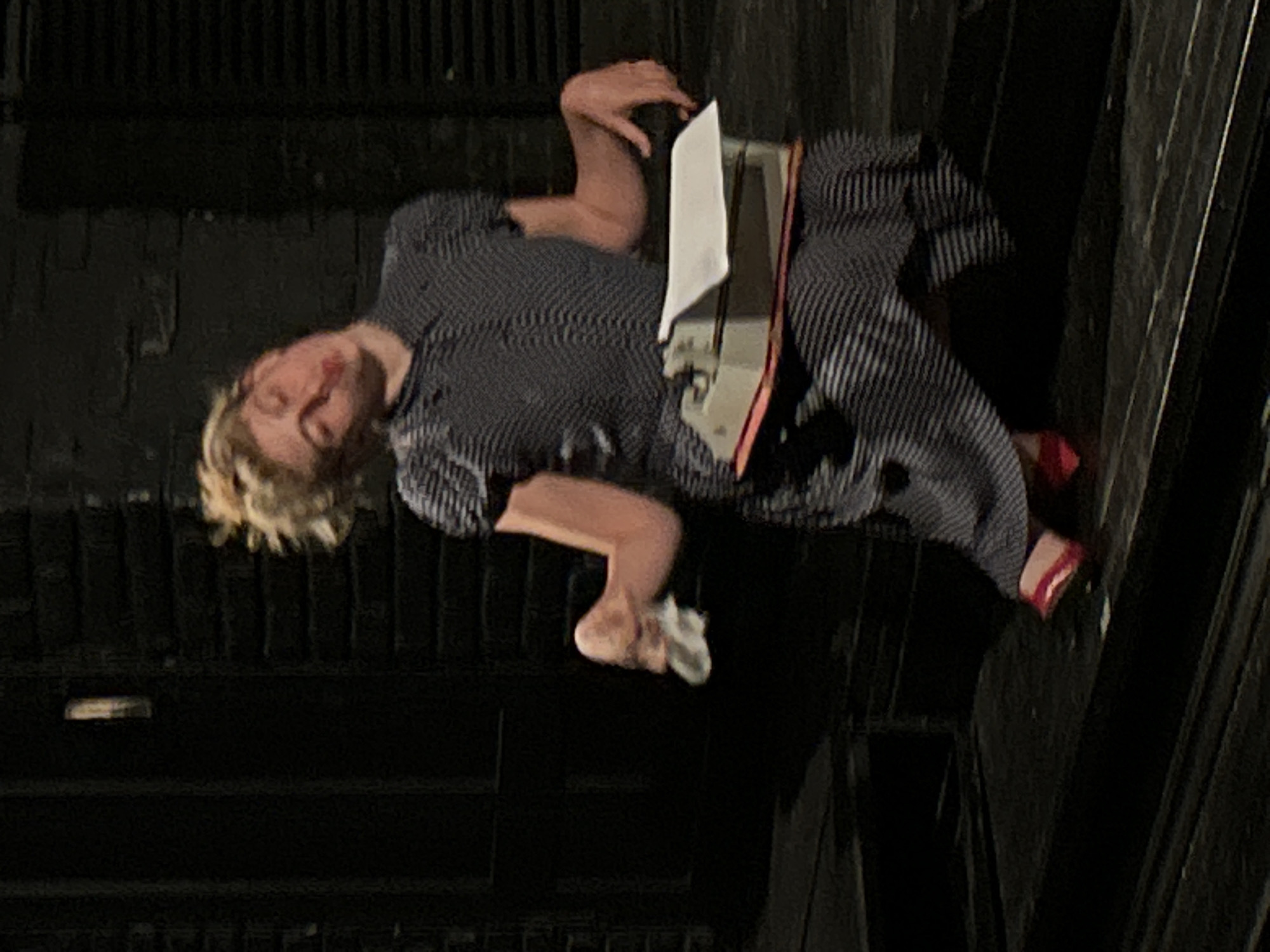 A medium shot depicts a person lying on their side atop what appears to be dark, possibly wooden slats. The individual, who seems to be female, is wearing a dark polka dot or patterned blouse, striped pants, and red heels. A metallic object, possibly a typewriter or similar machine, rests near the person's torso. A white piece of paper is situated near the machine. The background is dark and indistinct. The city and specific location are Unknown.