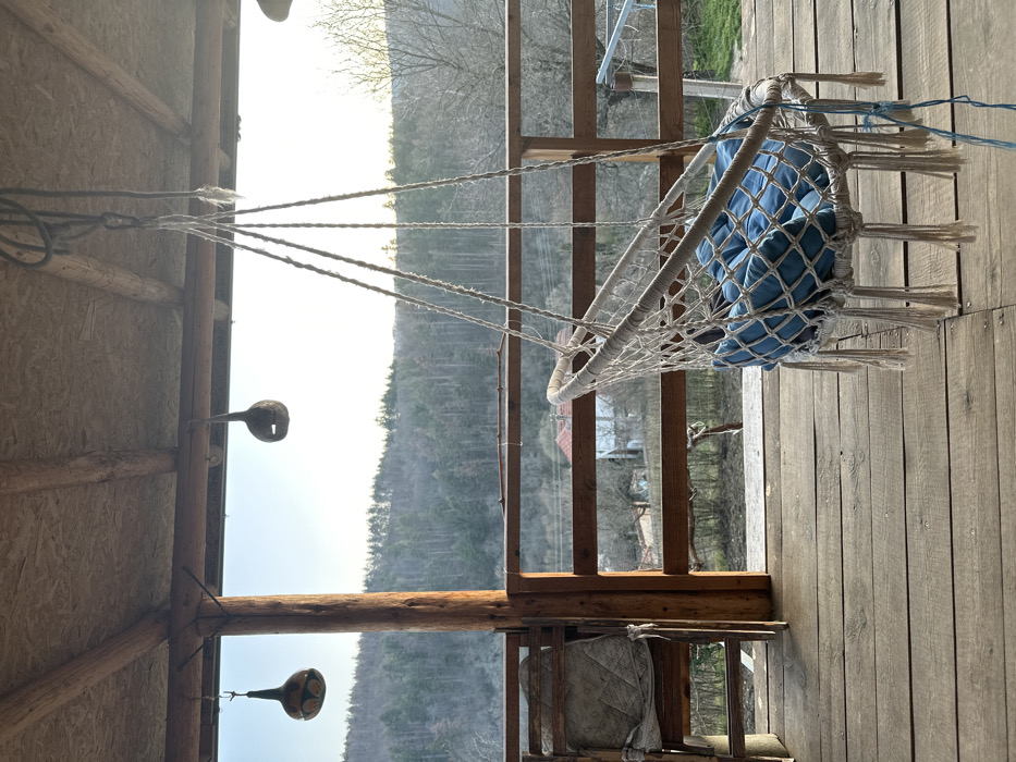 The image depicts a wooden porch or balcony overlooking a rural valley in Mrachenik, Bulgaria. The porch features a wooden floor composed of parallel planks, a thick wooden support column on the left, and a wooden railing spanning the mid-ground. Hanging from the wooden ceiling beams are two gourd-shaped decorative objects; the one on the left is painted with a pattern, while the one on the right is a dark, natural gourd with a cut-out. A white, netted hanging chair with blue quilted cushions rests on the right side of the frame, suspended by ropes from the ceiling. To the left, an empty wooden folding chair with a light-colored, patterned cushion is visible next to a small wooden table. Beyond the porch railing, a valley unfolds, revealing scattered houses with red roofs amidst trees in varying stages of foliage, suggesting early spring or late autumn. A dense forest of mostly evergreen trees covers the hills in the background under a bright, clear sky. No individuals are present in the scene.