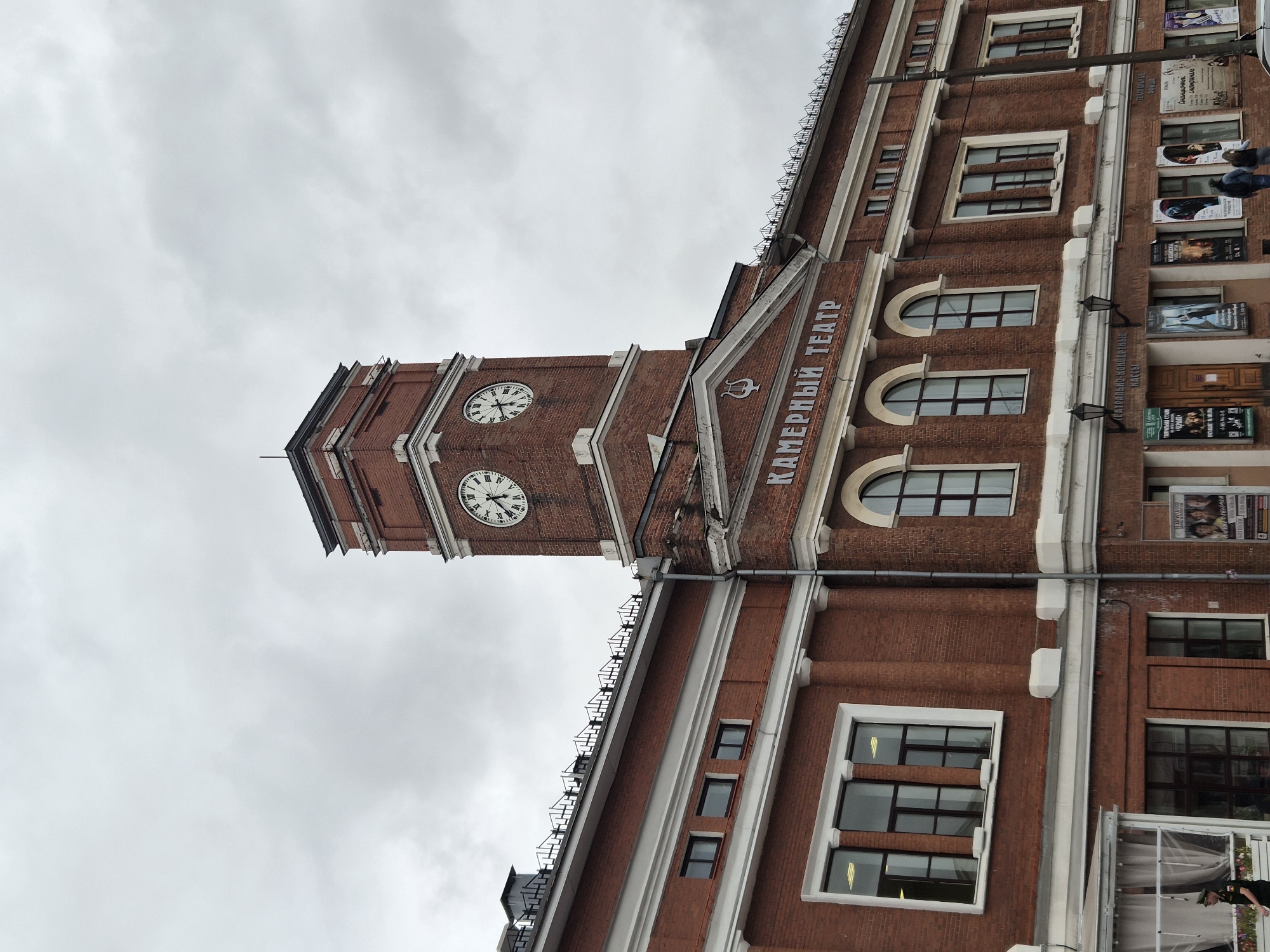 The image shows the exterior of the Cherepovets Chamber Theater (Камерный Театр) in Cherepovets, Russia. The building is constructed of red brick and features a prominent clock tower with two faces. The clock faces display Roman numerals, and the time appears to be approximately 3:35 on both. Above the main entrance, the words "КАМЕРНЫЙ ТЕАТР" are inscribed in white lettering. Several arched and rectangular windows are visible along the facade. Posters for theatrical performances are displayed on the lower level of the building. The sky is overcast. A person in a security uniform is standing to the left of the frame. Several other individuals are walking in front of the building on the right side.