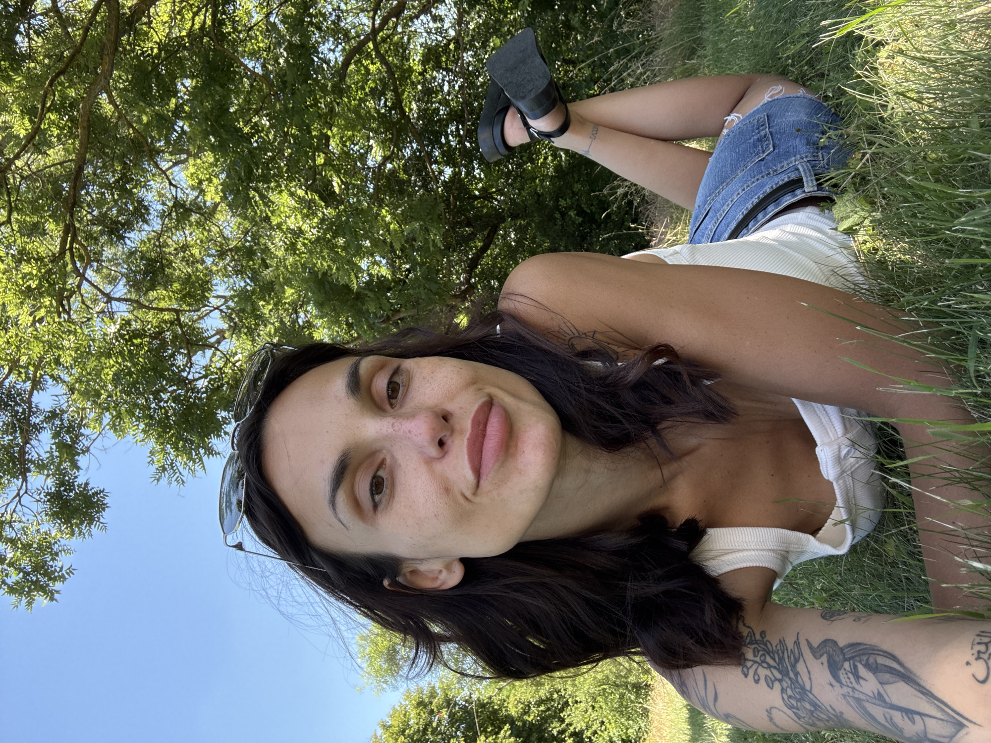 A woman is lying on her stomach in the grass in a park, with trees and a blue sky in the background. She is wearing a white tank top and denim shorts. Her legs are crossed and she is wearing black platform sandals. She has tattoos on her left arm and her right leg. Sunglasses are resting on her head. The location is the park in Pleven, Bulgaria.