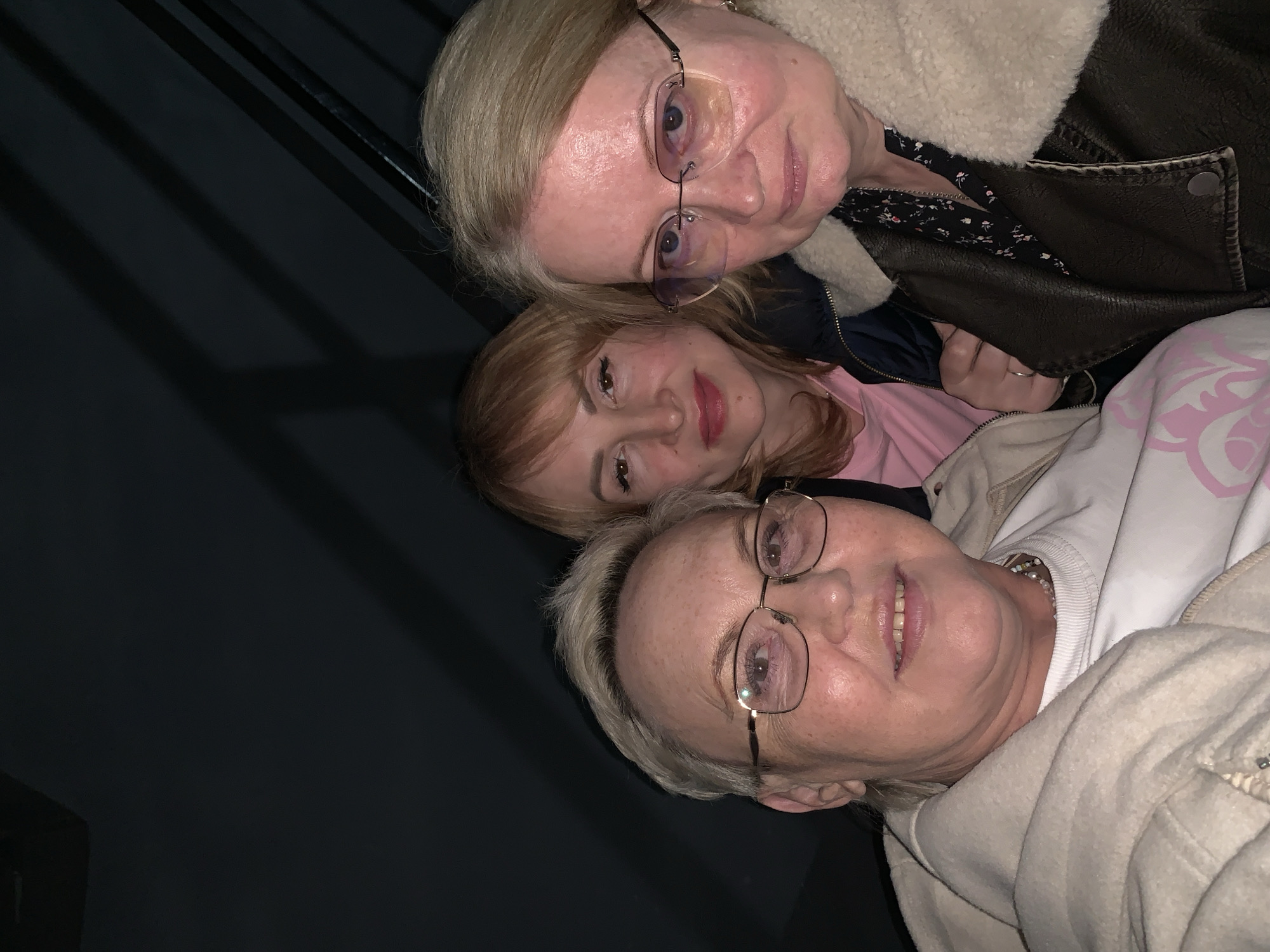 Three women are posing for a selfie-style photograph. The woman at the top has blonde hair, wears glasses, and is wearing a brown leather jacket with a light-colored fur collar and a floral-patterned top beneath. The woman in the middle has reddish-blonde hair and is wearing a pink top. The woman at the bottom has short, gray hair, wears glasses, and is wearing a light-colored jacket over a white shirt. The background is dark, suggesting an indoor location. The photograph may have been taken at an angle. The location is Chaikovsky, Russia.