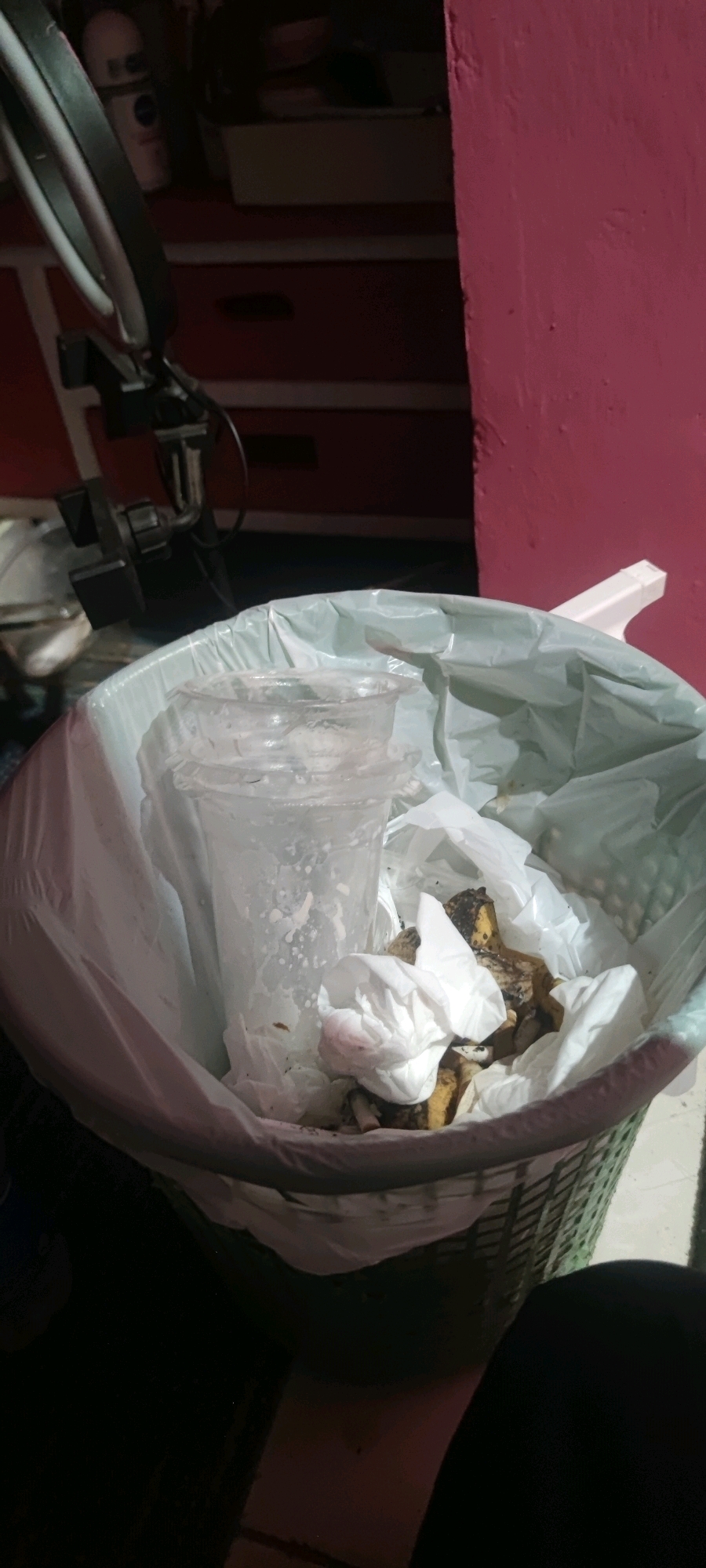 In Gambarsari, Indonesia, a close-up, low-angle shot captures a green woven trash can overflowing with refuse. Inside, a translucent plastic cup, likely containing residue, sits atop a white plastic liner. Scattered among the trash are used paper napkins and what appear to be discarded food scraps, possibly banana peels. In the background, a portion of a dresser with dark drawers is visible, along with a pink wall. A metallic lamp structure is also present, partially obscuring the dresser. The lighting is dim, with the primary light source appearing to originate from the foreground.