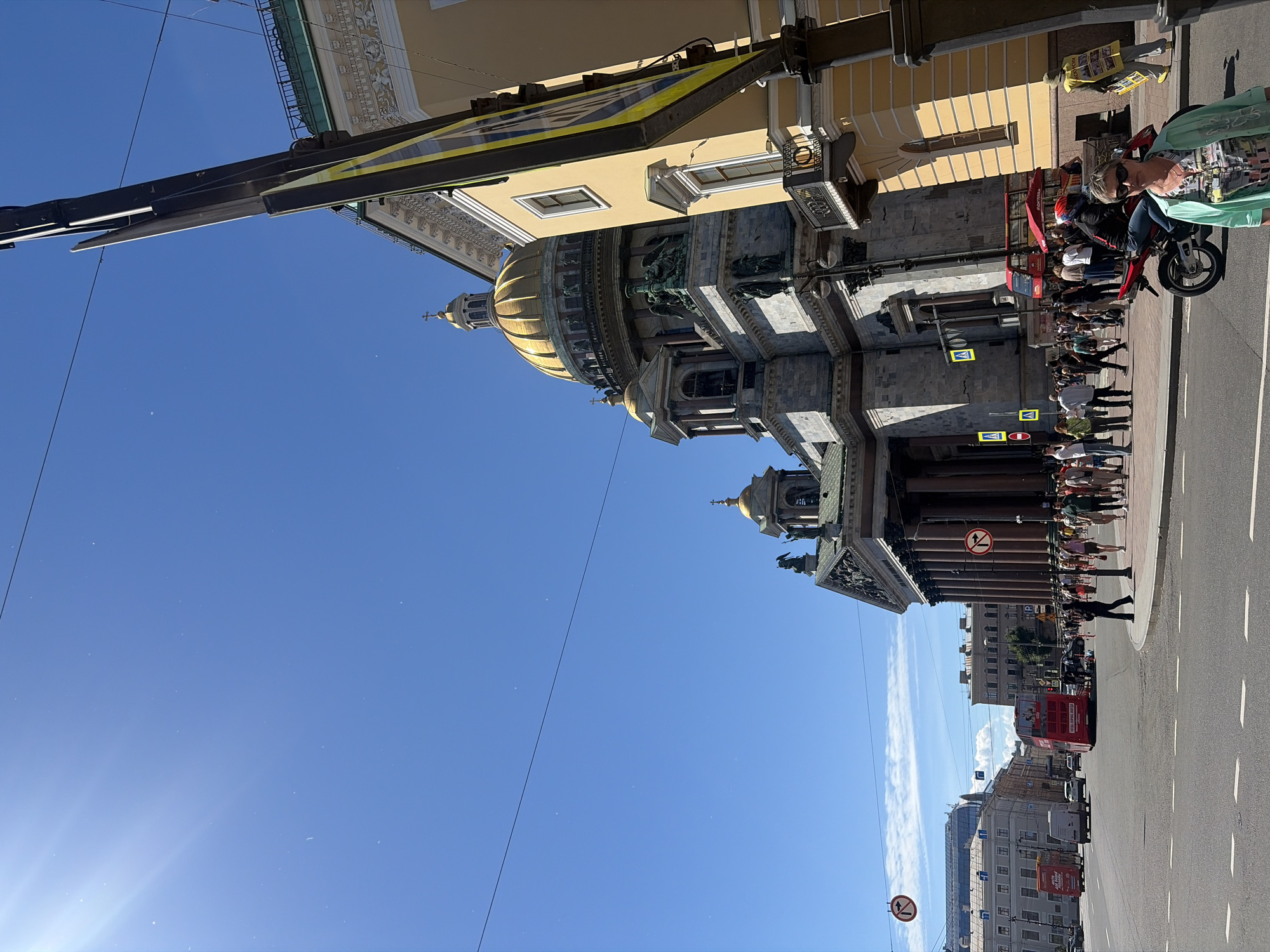 The image depicts a sunny day in Saint Petersburg, Russia. The scene is dominated by a grand architectural structure, likely a cathedral, with a prominent golden dome and intricate stonework. In the foreground, a woman on a red scooter is visible, wearing sunglasses and a light-colored garment with a colorful print. She appears to be looking at the camera. Behind her, a group of people are gathered, some standing and others walking. A red double-decker tour bus is also present on the street, suggesting a tourist area. Signs with international prohibition symbols are visible on poles. The sky is clear blue with a few wispy clouds. The lighting indicates it is daytime, likely midday given the bright sun. The overall mood is cheerful and active, with people enjoying the pleasant weather and the city's sights.