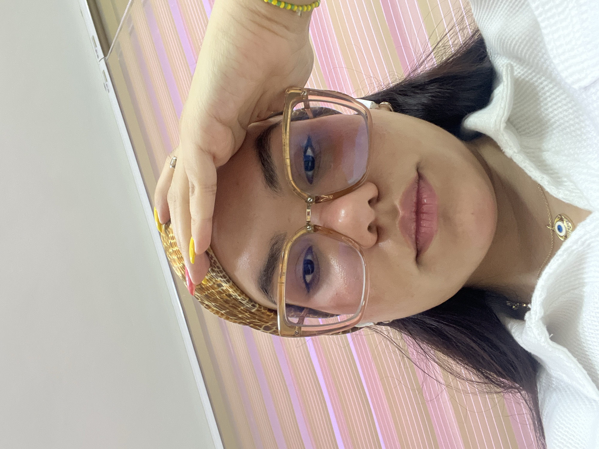A young woman with dark hair, wearing a leopard print headscarf, oversized clear-rimmed glasses, and a white collared shirt, is shown from the chest up. She is holding her right hand to her forehead. A gold necklace with a blue evil eye pendant is visible. The background consists of pink and beige striped blinds. The image is a close-up selfie. There is no indication of the location being La Asuncion, Venezuela.