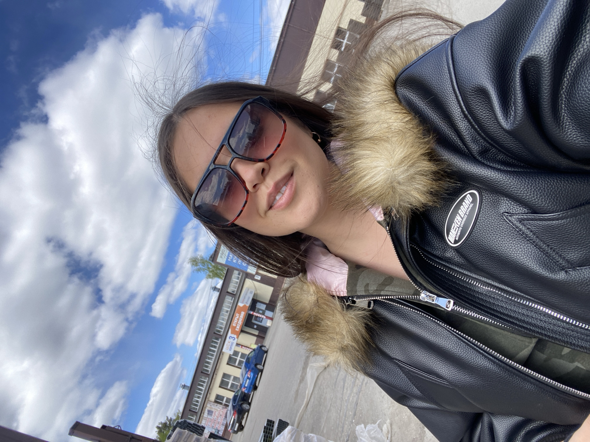A young woman wearing sunglasses and a black leather jacket with a fur collar is taking a selfie. The location is in Chaikovsky, Russia, as indicated by the Cyrillic lettering on nearby buildings. In the background, there are buildings, cars, and a partly cloudy sky.