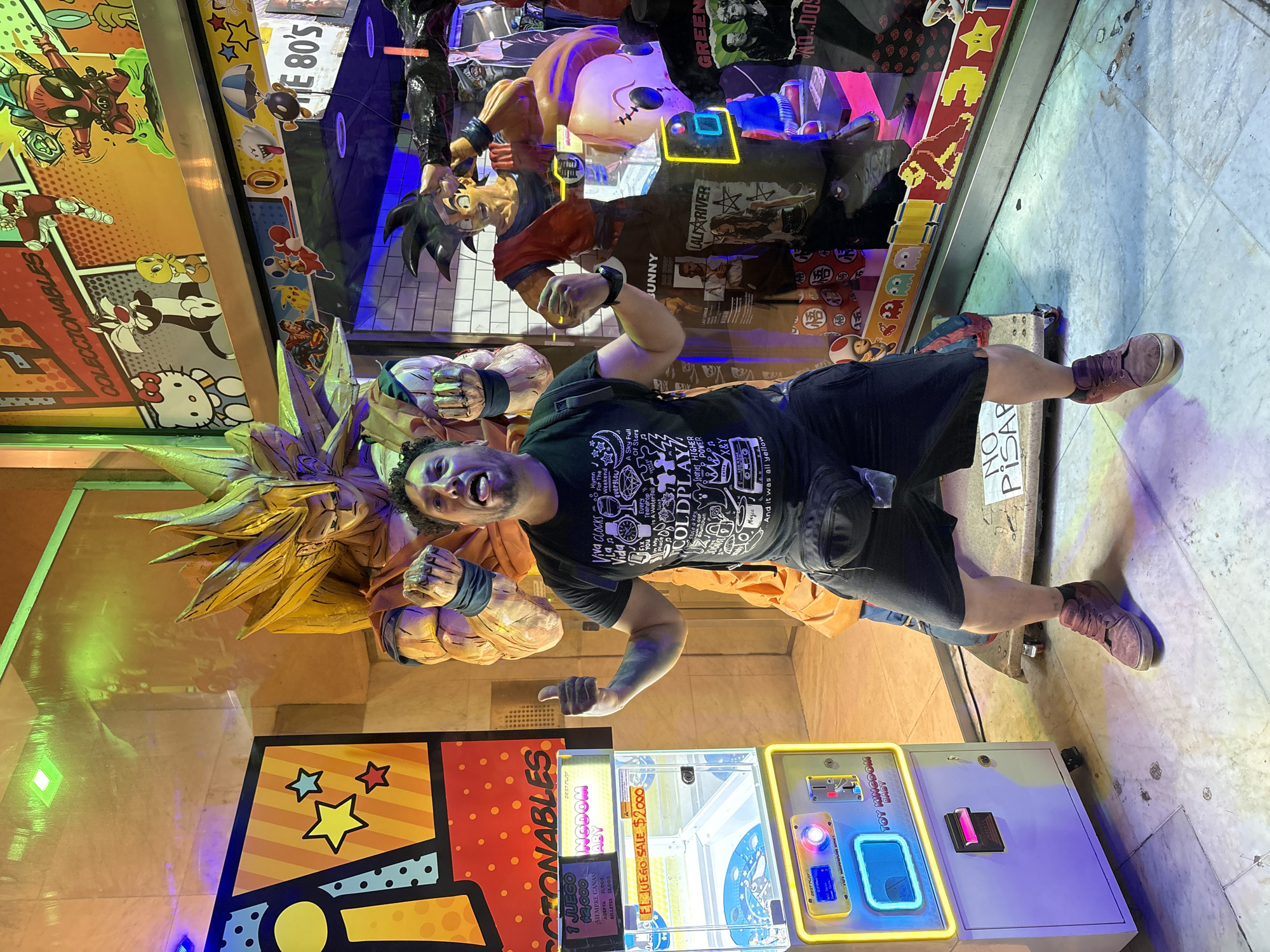 The scene is set in a commercial storefront entrance in Buenos Aires, Argentina, characterized by vibrant pop culture displays and artificial lighting.
An adult male, with short dark curly hair and a beard, is centrally positioned, facing the viewer. He wears a black t-shirt with white graphics, black shorts, and maroon sneakers, with a black fanny pack slung across his waist. He has a playful expression, sticking his tongue out, and both hands are raised with thumbs extended upwards.
Directly behind the man is a large, life-sized statue of Super Saiyan Goku from Dragon Ball Z, with yellow spiky hair and an orange gi, standing on a wheeled base. To the man's left is a clear plastic arcade claw machine, labeled "TOY KINGDOM BABY," featuring neon yellow and blue lights. Its display shows "1 JUEGO $2.000" (1 Game $2,000) and "SIEMPRE GANAS" (Always Win).
The background is a brightly illuminated retail display window, adorned with various cartoon and comic book characters in a comic panel style, including Hello Kitty, Sylvester the Cat, Tweety Bird, Deadpool, Superman, Pikachu, Mario, and Donkey Kong. A sign above reads "COLECCIONABLES" (Collectibles), and another indicates "THE 80'S". Inside the display, a standard Goku statue (red gi) and other merchandise, including t-shirts (one "GREEN DAY"), are visible. A white sign reading "NO PISAR" (Do Not Step) is on the light-colored, tiled floor near the man's feet.