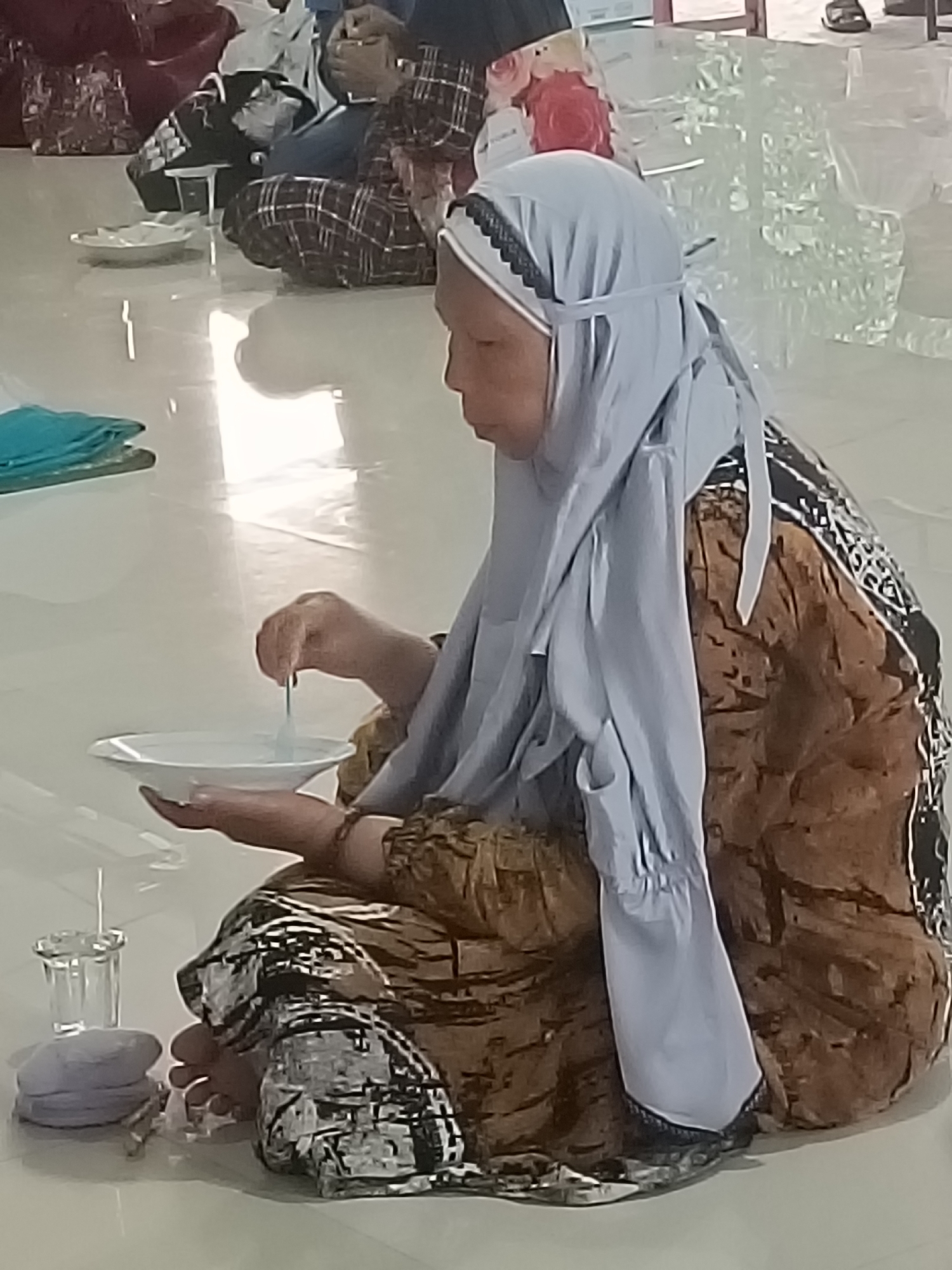 A woman wearing a light blue hijab and a brown and black patterned dress is sitting on the floor, holding a white bowl and stirring its contents with a spoon. She appears to be eating or preparing food. The floor is white and reflects light, suggesting an indoor setting, possibly a community hall or a religious gathering space in Kediri City, Indonesia. Other individuals, partially visible, are also seated in the background, some engaged with their phones. The overall scene is casual and domestic.