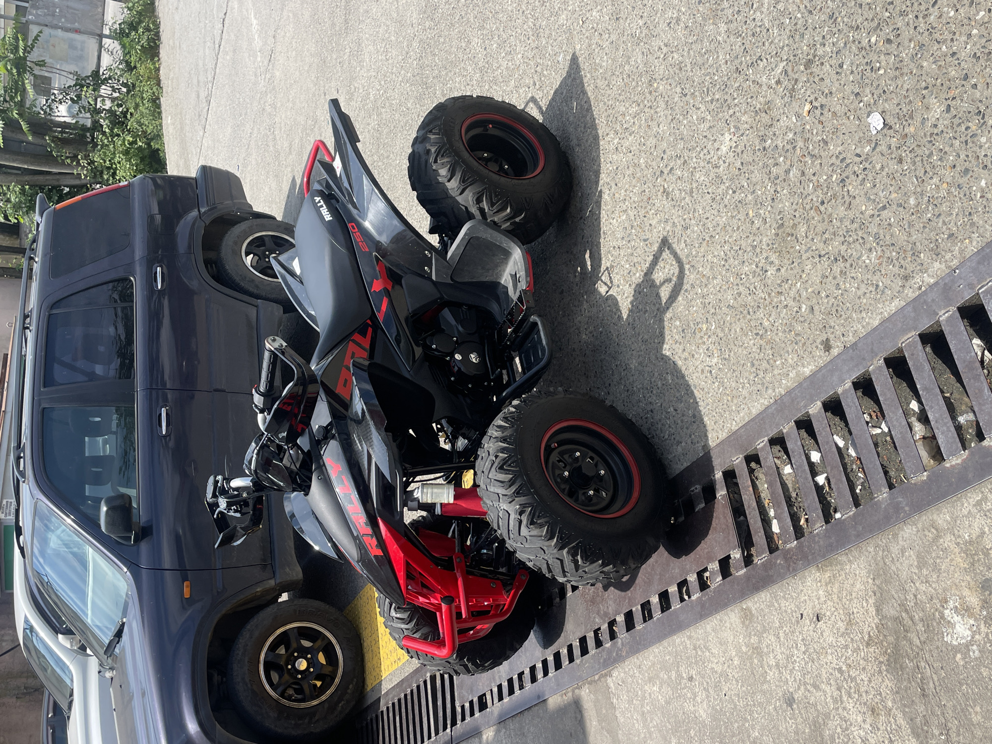A black and red ATV is parked on a concrete surface next to a dark-colored SUV. The ATV has knobby tires with red rims and a red front bumper. The SUV appears to be a Honda CR-V. A metal grate runs along the ground in front of the ATV. The scene is outdoors, with trees and buildings visible in the background. There are no people in the image.