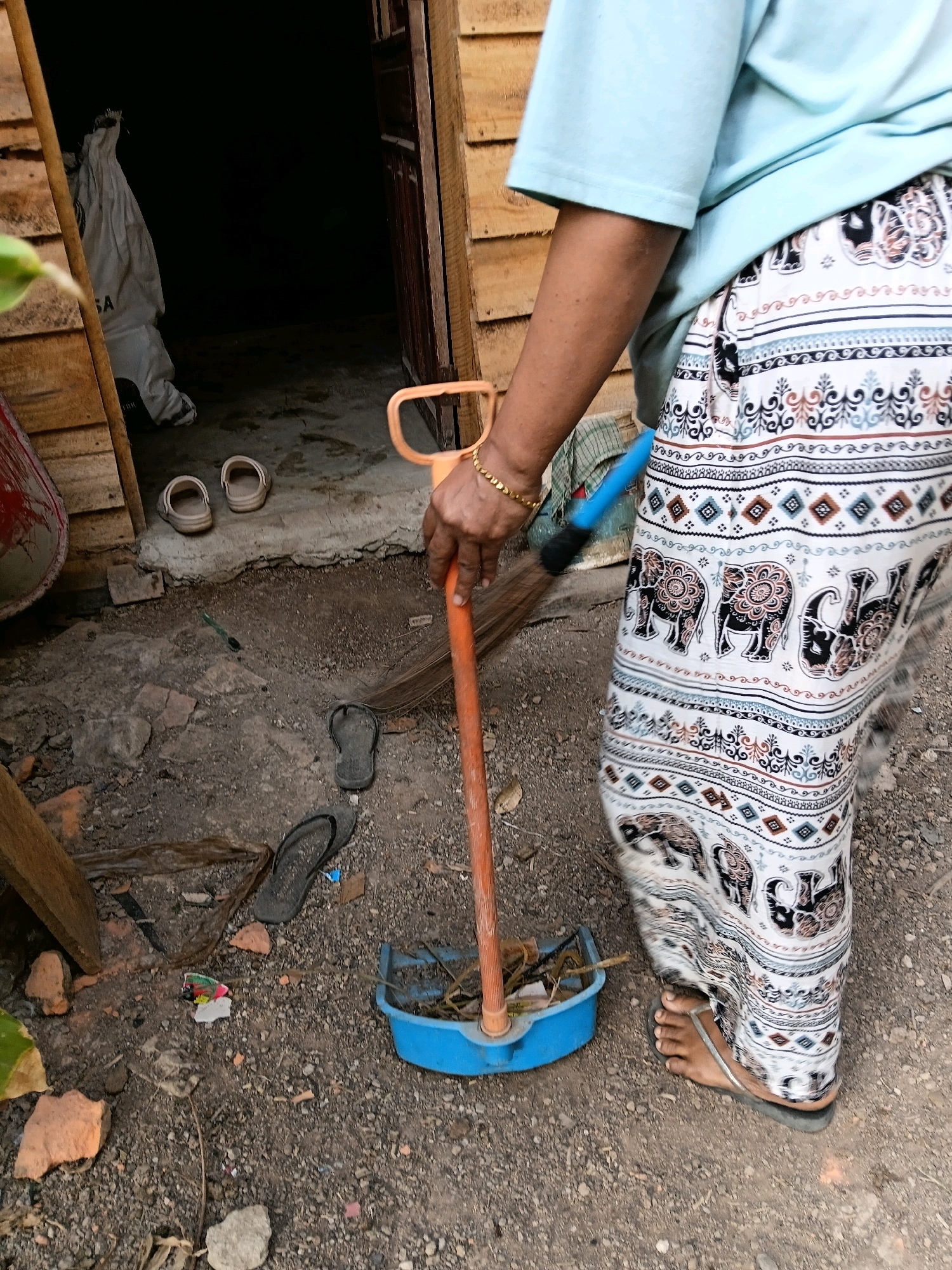 An individual, wearing a light blue t-shirt and a patterned skirt with elephants, is standing on a dirt ground outside a wooden structure. The individual is holding an orange-handled broom and is in the process of sweeping debris into a blue dustpan. Several pairs of flip-flops and clogs are scattered on the ground around the individual. In the background, an open doorway leads into a dark interior space, where a large white bag is visible. The scene suggests a domestic or outdoor working environment in Margo Rukun, Indonesia.