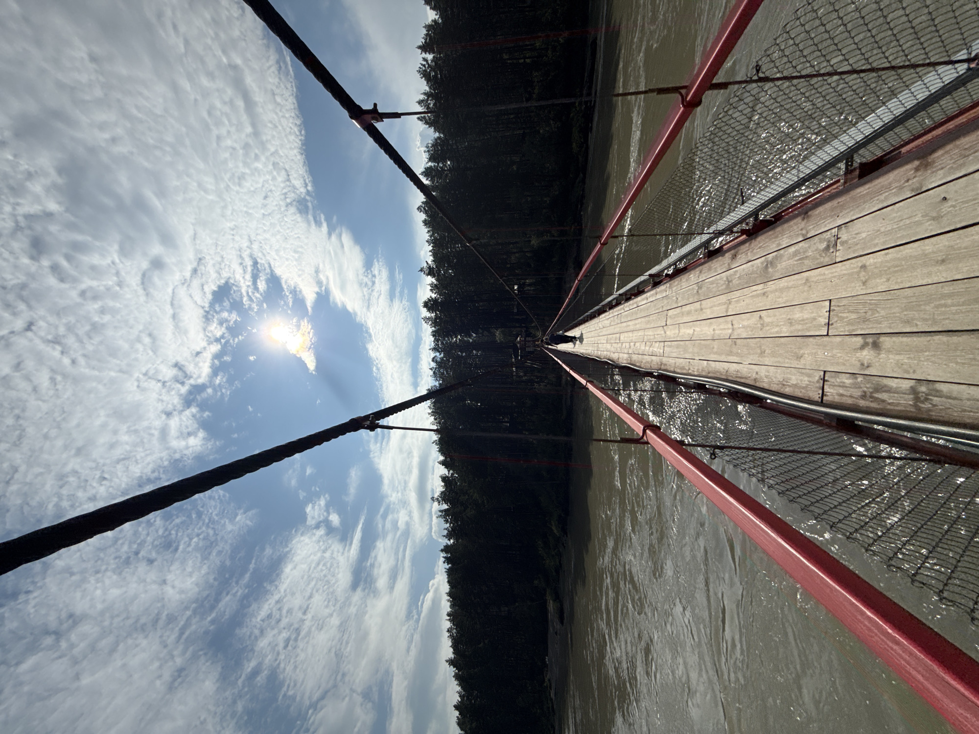 The image depicts a person walking across a wooden suspension bridge over a river. The bridge has red railings and is supported by thick cables. A dense forest of dark green trees lines the opposite bank of the river. The sky is blue with scattered white clouds, and the sun is visible, casting a bright glare. The river appears to be flowing with some turbulence. This scene is characteristic of the natural scenery found in and around Barangol, Russia, likely a tourist area given the presence of a pedestrian bridge over a river.