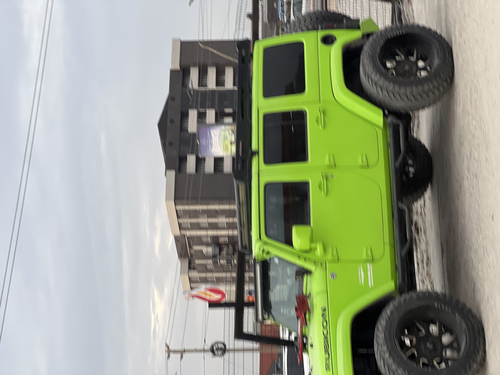 The image, taken in Igboefon, Nigeria, showcases a striking lime green Jeep Wrangler, identified by the visible "RUBICON" text on its side. The vehicle appears heavily modified for off-road use, featuring large, rugged tires on black multi-spoke rims, robust side steps, and a roof rack. A spare tire is mounted on the rear, and a red tool, possibly an axe or shovel, is attached to the front fender area.
The Jeep is positioned on what seems to be a paved or concrete surface. In the background, a multi-story building with dark paneling and lighter-colored architectural elements stands prominently. A sign or billboard is affixed to this building, though its details are not clearly legible. Another building is partially visible further in the distance. Overhead, power lines crisscross against an overcast, cloudy sky, indicating a daytime setting. No people are visible in the scene, and the Jeep appears to be stationary or moving slowly within an urban or semi-urban environment.