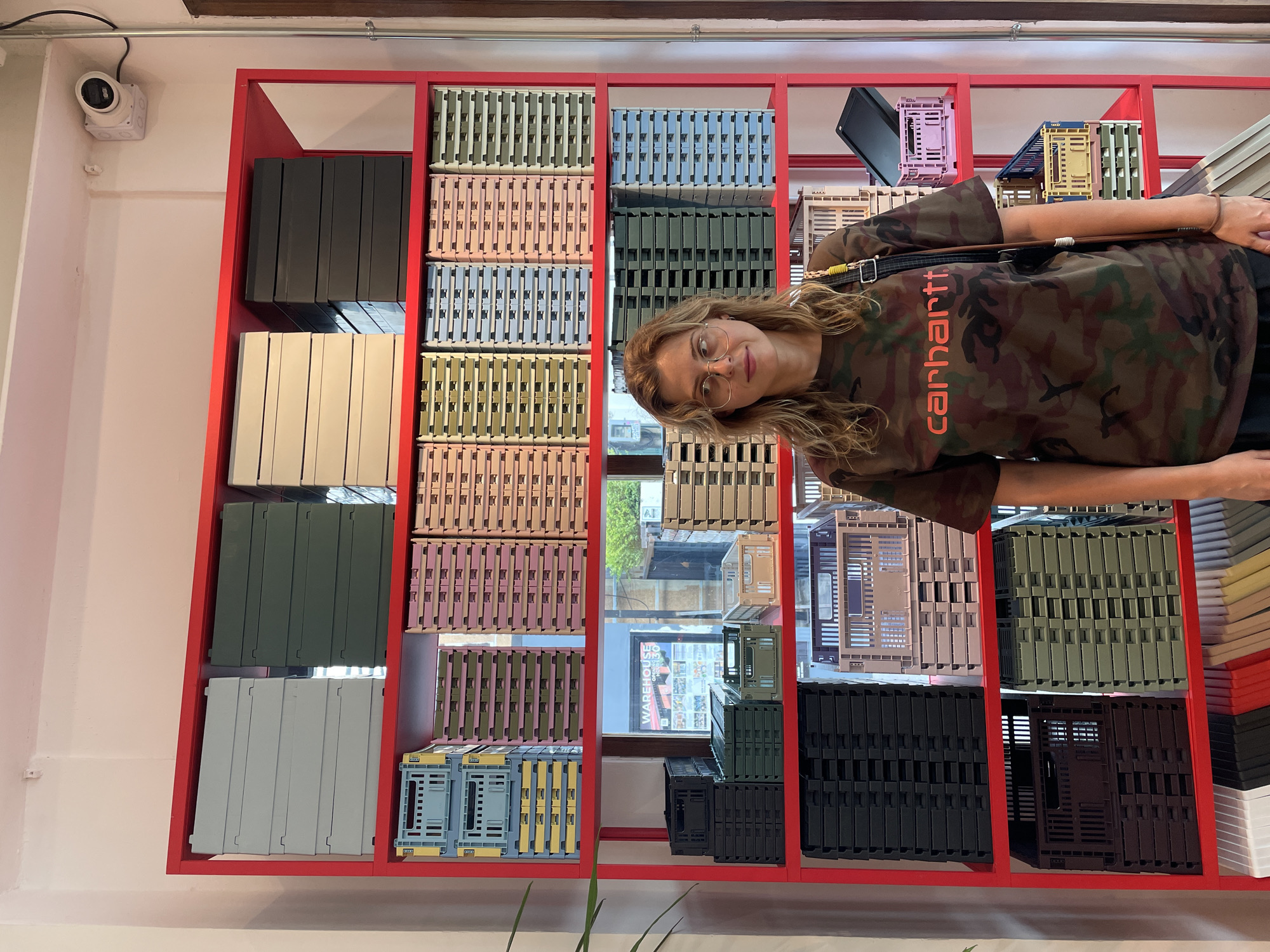 The image shows an indoor scene in Bangkok, Thailand. A woman wearing glasses and a Carhartt camo-patterned shirt is standing in front of a red shelving unit filled with colorful plastic crates. The shelving unit appears to be part of a store display. Outside the store window, the word "WAREHOUSE" is visible. A security camera is mounted on the wall near the top left corner of the frame.