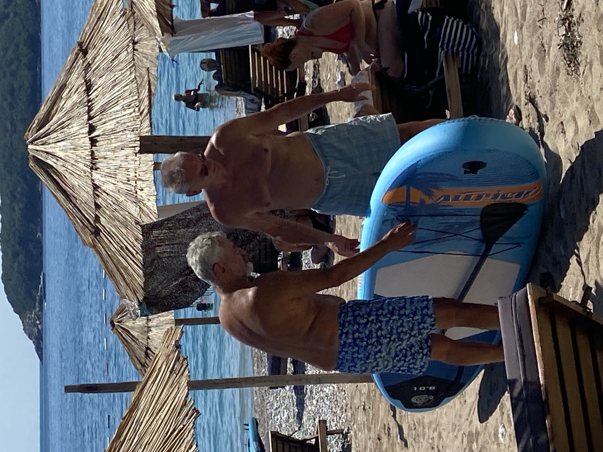 In Prijevor, Montenegro, on a sunny beach, two shirtless men stand on the sand. The man on the left wears blue patterned swim trunks and leans over a blue inflatable paddleboard, touching it with his right hand. The man on the right wears light blue plaid swim trunks and stands looking forward. Behind them, the turquoise sea meets a tree-covered hill. Several straw umbrellas provide shade on the sandy shore, where beach chairs and other beachgoers are visible in the background. The scene captures a relaxed summer atmosphere.