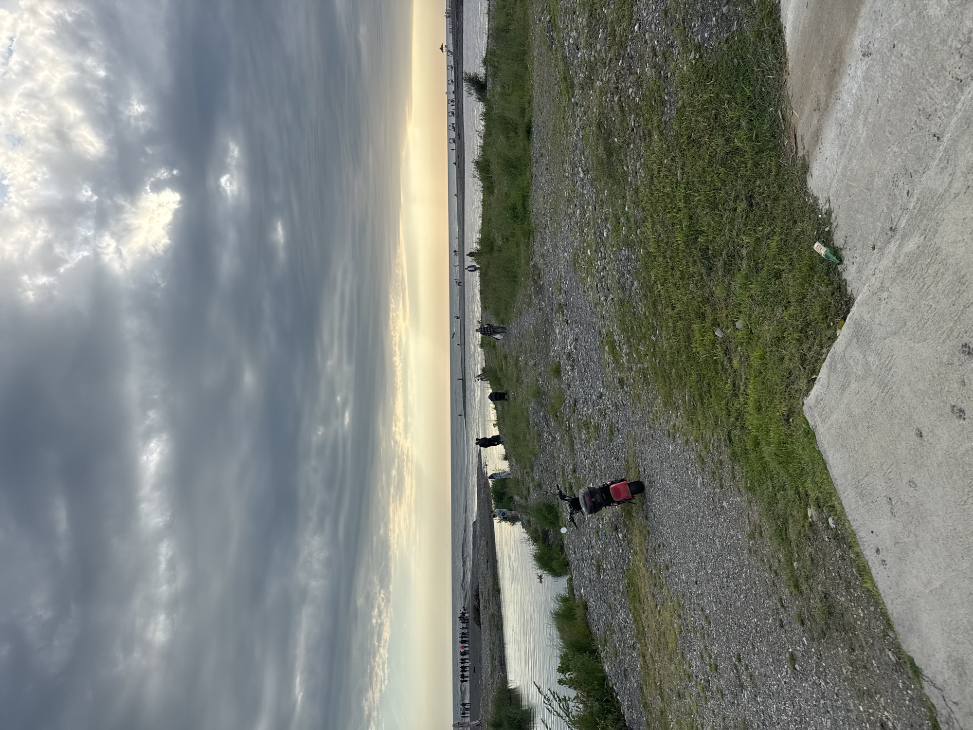 The image shows a coastal scene in Sirius, Russia. A wide expanse of water is visible, stretching towards the horizon under a cloudy sky with hints of sunset. Along the shoreline, there is a pebbled area with patches of grass. Several people are walking along the beach, and a red motorbike is parked near the water's edge. A concrete pathway or wall is visible on the right side of the frame, with some litter nearby. The overall impression is of a calm evening near the sea.