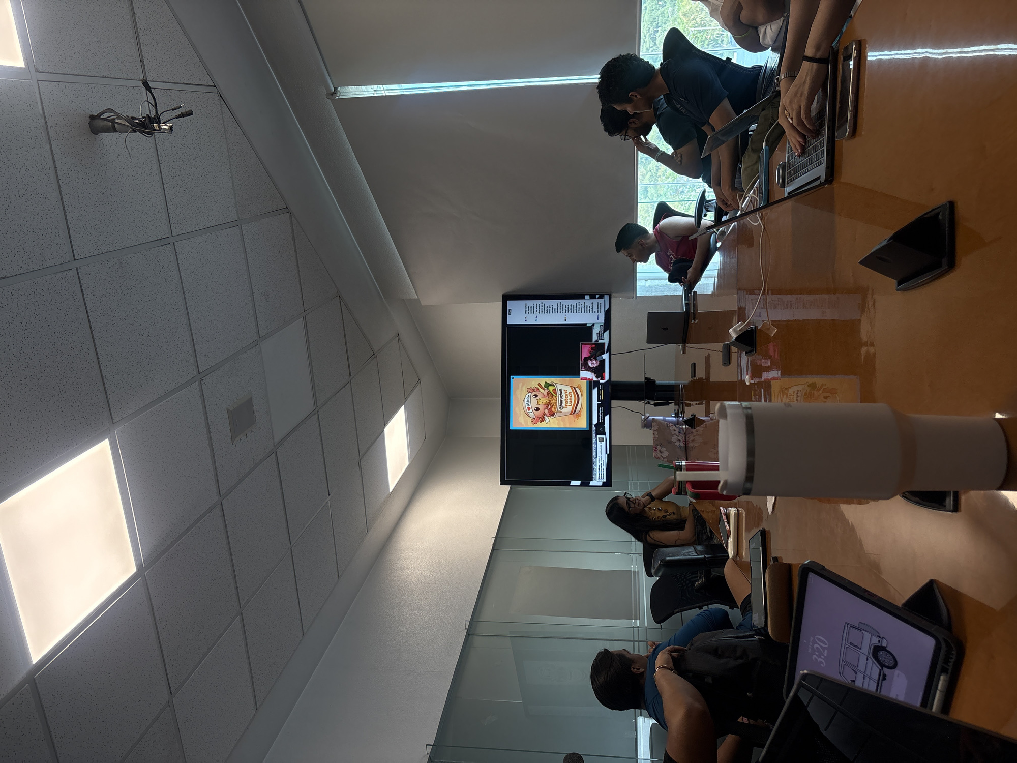 A brightly lit conference room appears to be in a commercial office setting. There is a large conference table surrounded by people working on laptops and other electronic devices. A large screen is visible on the wall, displaying what seems to be a video conference call. The room has a drop ceiling with fluorescent lighting, and a wall of glass separates the room from an adjoining space. Based on the logo seen on the screen, the location is likely in Puebla, Mexico.