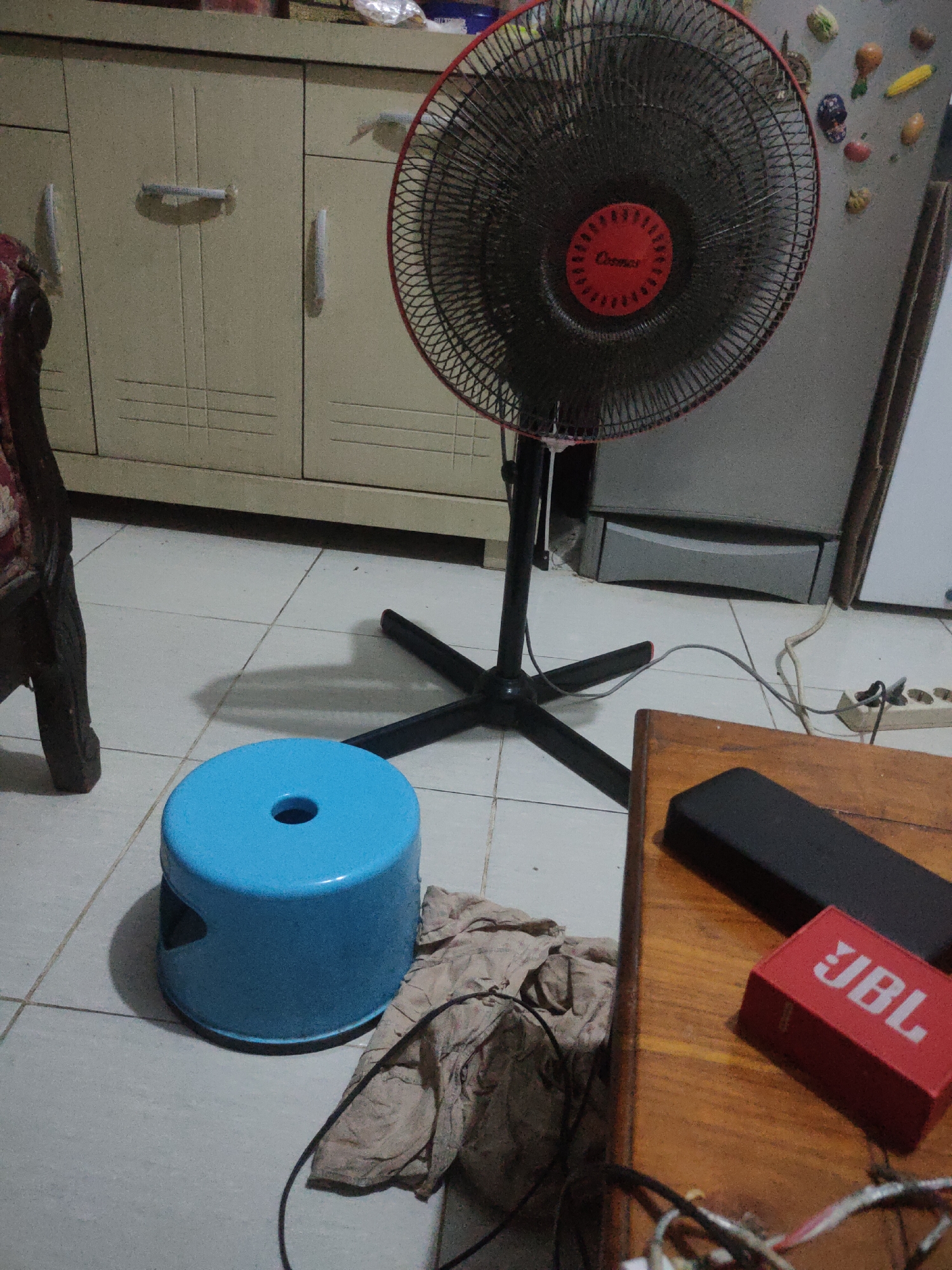 The image depicts an indoor scene, likely a room in South Jakarta, Indonesia. In the foreground, a blue plastic stool with a hole in the center sits on a tiled floor. Next to the stool is a crumpled piece of light brown fabric. To the right, a portion of a wooden table is visible, with a black rectangular object and a small red JBL speaker on its surface. In the mid-ground, a black standing fan with a red center emblem is positioned. The background shows a beige cabinet with horizontal and vertical lines suggesting drawers or doors, and a wall with decorative magnets. A white power strip with plugs is also visible near the fan. The overall environment suggests a residential or storage area.