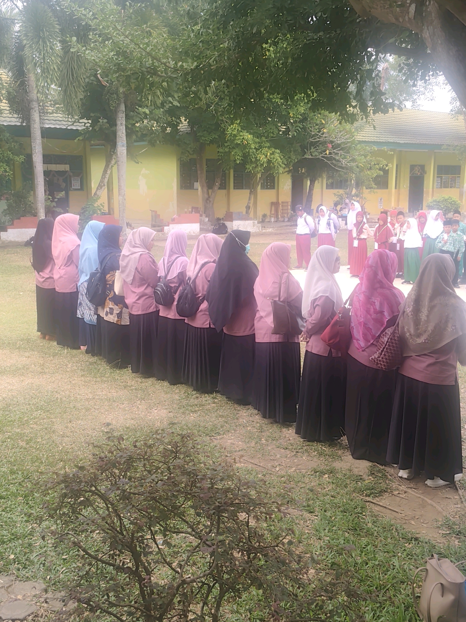 The image shows a group of young people, primarily girls, standing in a line outdoors. They appear to be students, as some are wearing uniforms consisting of long skirts and blouses, and many are wearing hijabs. The setting looks like a schoolyard, with a building in the background that has yellow walls and a green roof. There are trees and grass in the foreground and middle ground, suggesting an outdoor, possibly tropical environment. Some students in the background are interacting, and others are standing by themselves. The overall mood seems to be casual and possibly indicative of a school event or gathering. The image was taken in Pematang Reba, Indonesia.