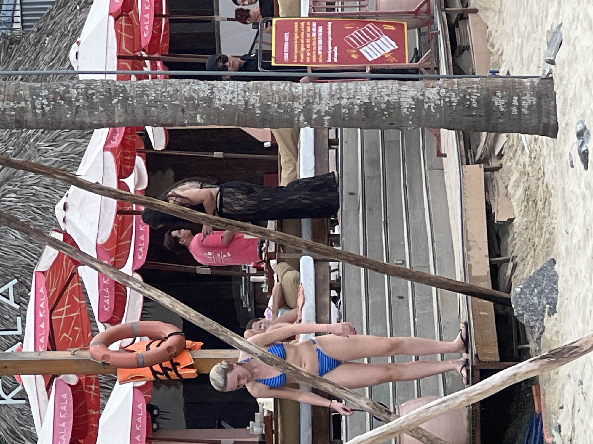 The image shows a beachside establishment in Đà Nẵng, Vietnam, possibly a restaurant or bar. In the foreground, a woman in a blue and black striped bikini stands on a wooden deck, looking down. Behind her, a person in a bright pink t-shirt stands and gestures with their hand. Further back, a person in a black lace dress is visible. Various beach umbrellas with the logo "KALA KALA" are set up. A life vest and a life preserver are hanging from a wooden post. A palm tree trunk is prominent in the right side of the image, and a red sign with white text and illustrations is mounted on a stand next to it. The scene is set on a sandy beach with wooden structures and what appears to be outdoor seating.