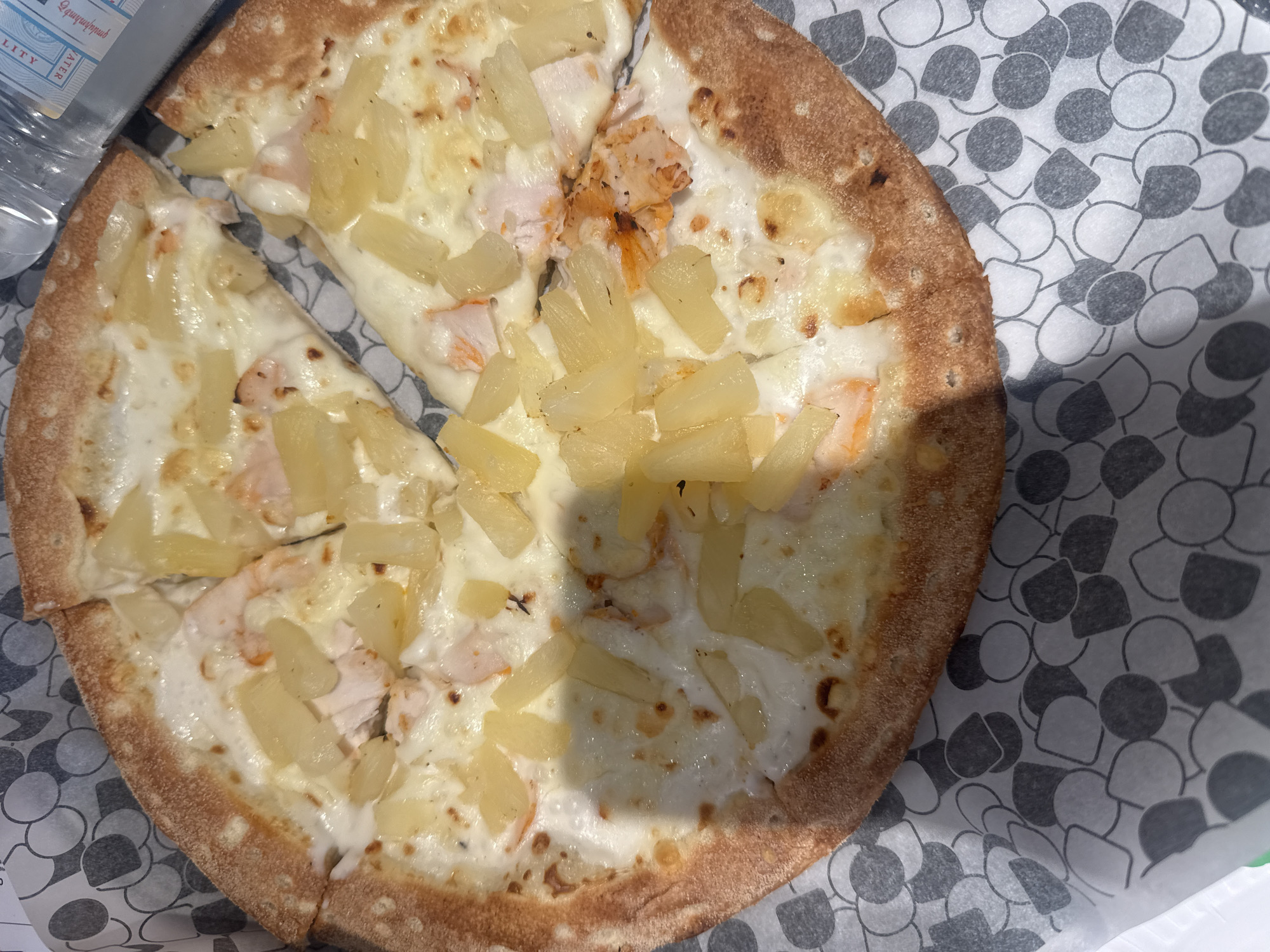 The image shows a partially eaten pizza topped with white sauce, chicken, and pineapple chunks on a patterned paper plate. The pizza is served in an outdoor setting, possibly a cafe or restaurant. The location is Yerevan, Armenia. A bottle is partially visible in the upper-left corner.