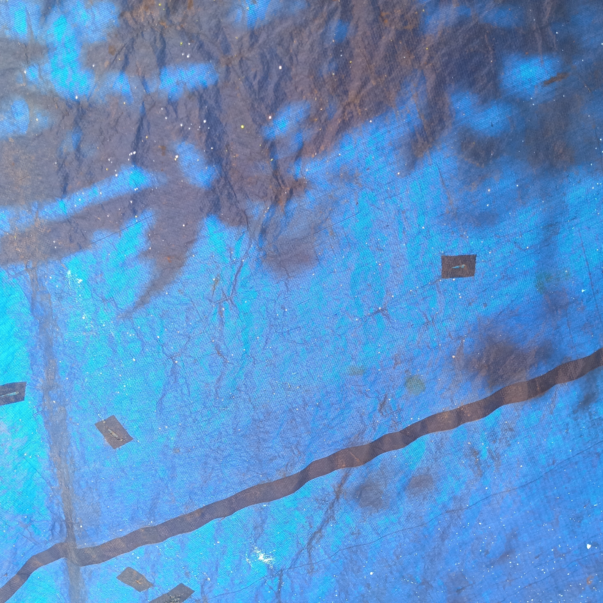The image displays a close-up view of a blue tarp, likely outdoors, as evidenced by the dappled light and shadows on its surface. The tarp has a woven texture and is creased and wrinkled, indicating it is flexible and has been manipulated. There are several dark rectangular patches affixed to the tarp, possibly for reinforcement or repair. The tarp is predominantly blue with some darker, shadowy areas and scattered white specks that could be dust or debris. The overall impression is one of a utilitarian object that has been exposed to the elements. The location is specified as Tumampua, Indonesia.