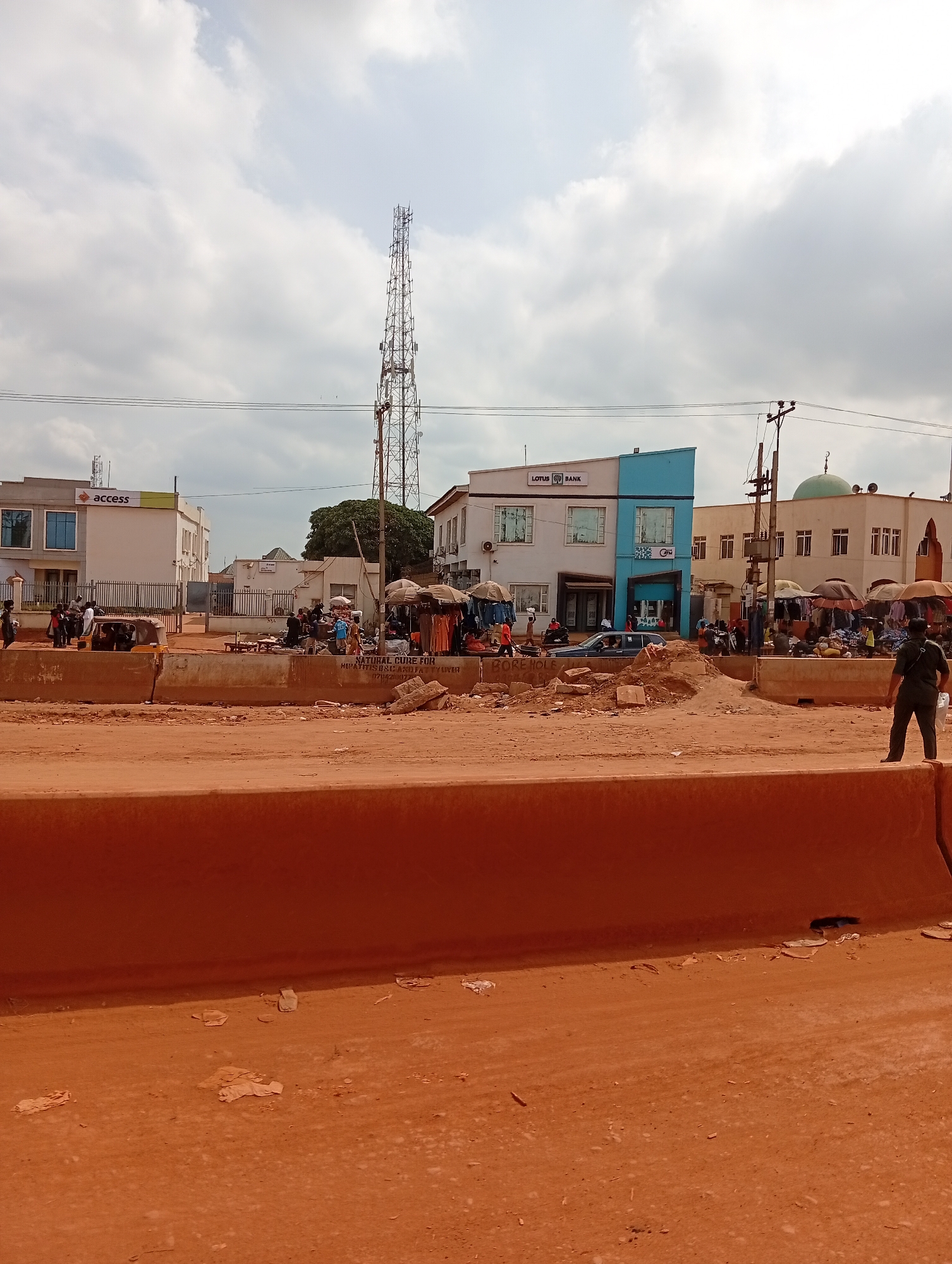 The image depicts a street scene in Ado, Nigeria, with a red-dirt road in the foreground partially blocked by a concrete barrier. Beyond the barrier, a bustling marketplace is visible. Various stalls with vendors and customers are scattered along the street. Buildings flank the marketplace, including what appear to be bank branches (ACCESS and LOTUS BANK) and a mosque with a green dome. A tall communications tower rises in the background. A man stands on the right side of the road. The sky is overcast.