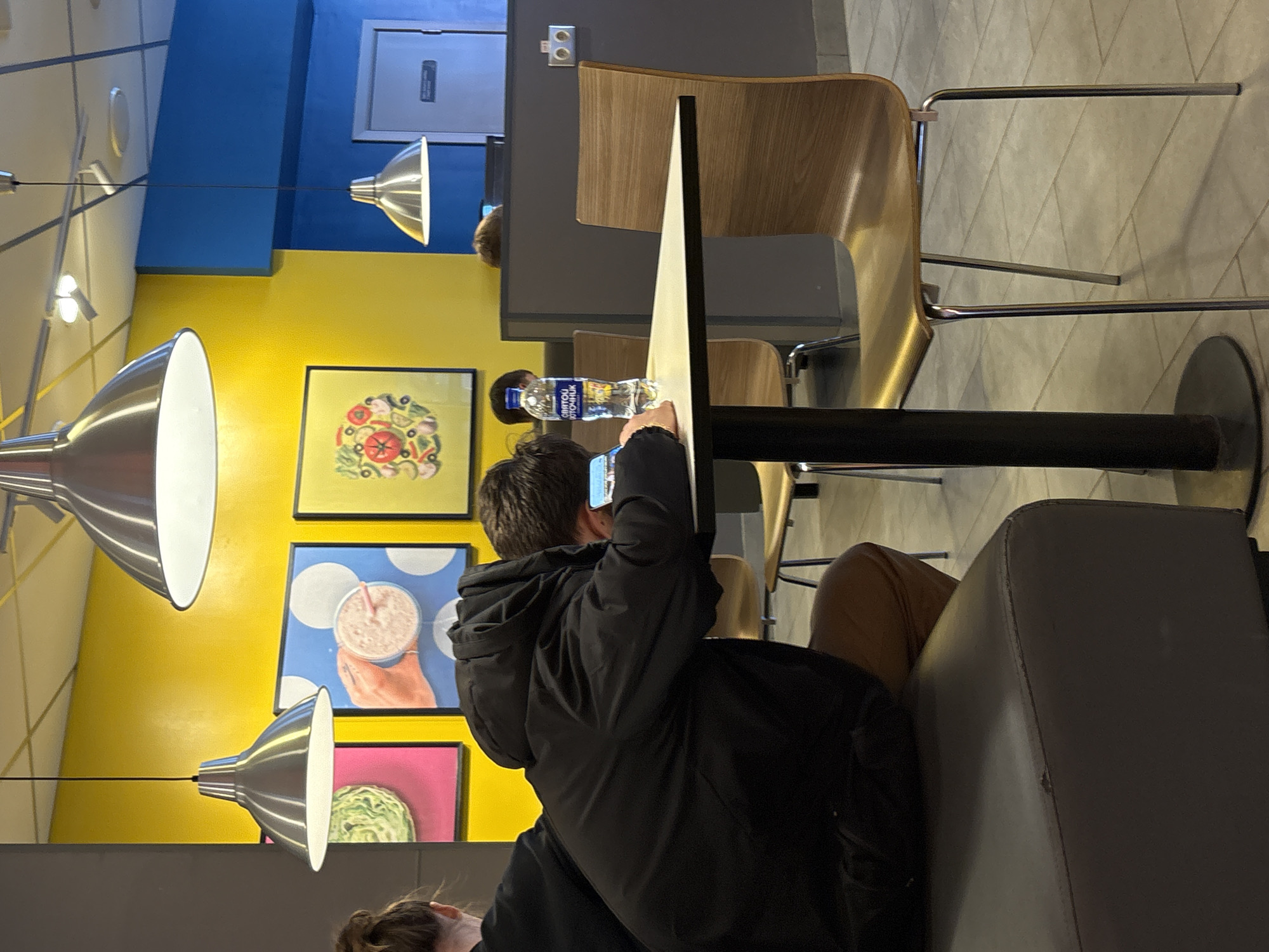 The image captures an indoor scene, likely within a modern cafe or casual dining establishment in Votkinsk, Russia. The walls are distinctly painted in alternating sections of bright yellow and blue. The ceiling features several contemporary pendant lights with metallic, conical shades, illuminating the space.
In the foreground, a person is seated at a dark gray booth, facing away from the camera. They are wearing a black puffer jacket and are actively holding up a smartphone, seemingly taking a picture or video. Only the top of their head and a dark hair bun of another person are partially visible in the bottom left corner. Further back, another individual with brown hair is seated at a table, near a plastic water bottle.
The tables feature light wooden chairs with metal frames and dark gray tabletops. On the bright yellow wall, three framed pictures are prominently displayed: the top one shows a colorful, circular food item resembling a pizza or salad; the middle one depicts a hand holding a milkshake with a straw against a blue polka-dot background; and the bottom one features a green, textured object, possibly a vegetable like lettuce or cabbage, against a vibrant pink background.
A plastic water bottle with a blue label, bearing Cyrillic text, rests on a table in the mid-ground. The visible text on the bottle reads "МИНУСИНСКАЯ" (Minusinskaya) and "ПРИРОДНАЯ ПИТЬЕВАЯ ВОДА" (Natural Drinking Water). The floor has light-colored tiles. An emergency exit sign or panel is visible on the blue wall section in the background. The overall atmosphere suggests a casual and brightly decorated dining environment.