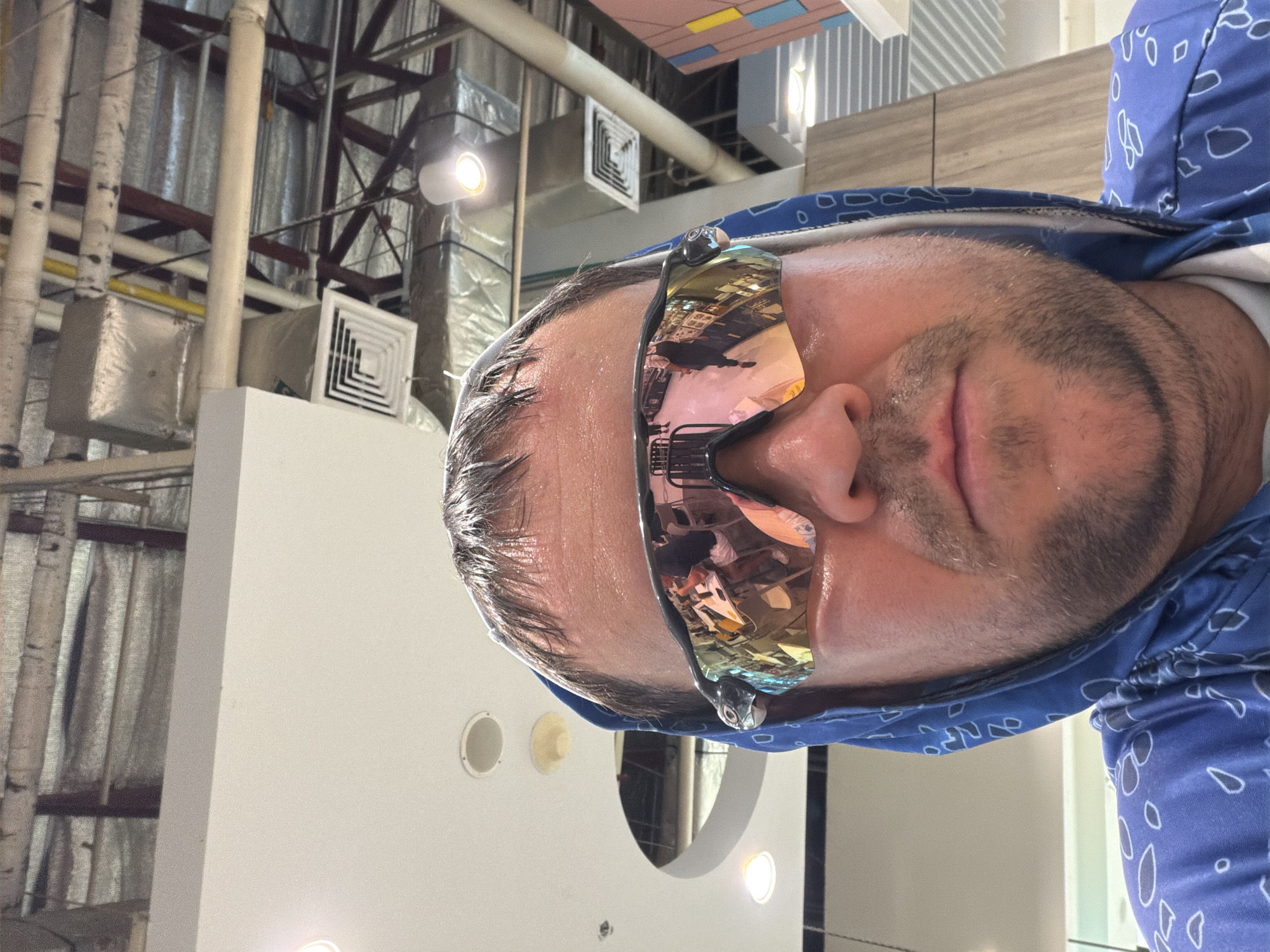 In a selfie taken in Ban Rang Rak, Thailand, a person wearing a blue hooded garment with a white paisley pattern is shown from the chest up. They are wearing reflective sunglasses, which show a distorted reflection of a busy interior space, possibly a shopping mall or food court. The background features exposed industrial ceiling elements, including ductwork, lighting fixtures, and structural beams. The person's face appears to be sweating, suggesting warm weather or physical exertion. The lighting in the scene is artificial and somewhat harsh, contributing to the overall high-contrast appearance of the image.