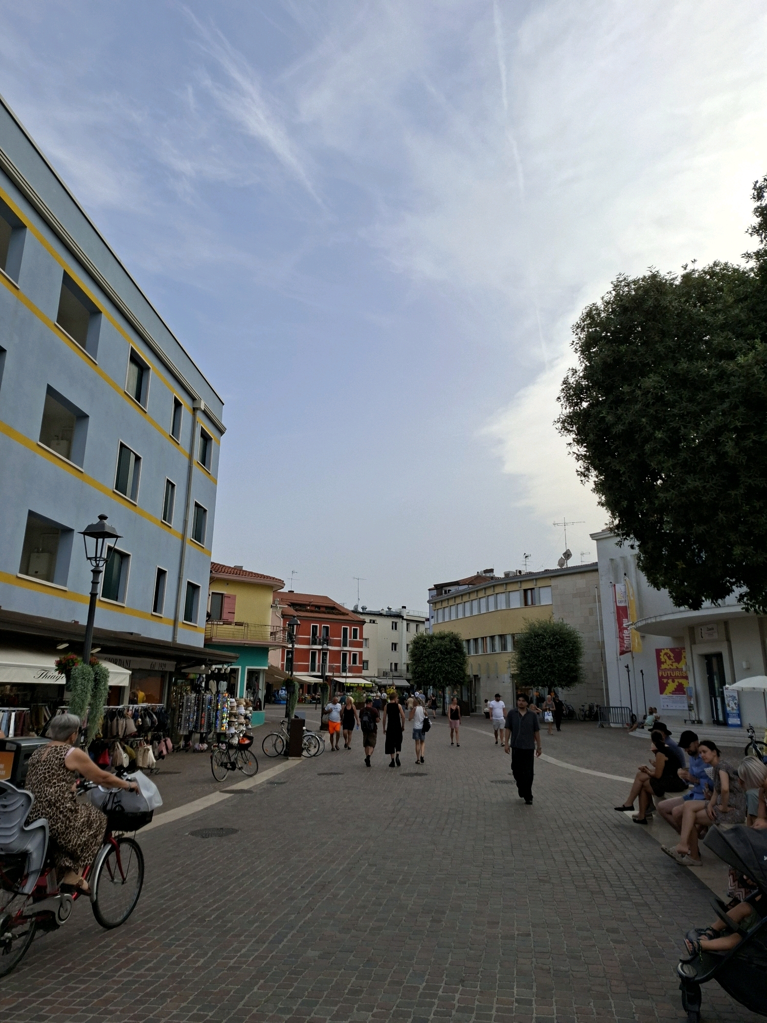 The image depicts a pedestrian street in Caorle, Italy, with buildings lining both sides. The sky is clear with some wispy clouds. On the left, a blue building with yellow accents and numerous windows dominates the foreground. A lamppost stands in front of it. Shops are visible at the ground level of the building. Several people are walking and cycling on the paved street. A woman in a leopard-print dress rides a bicycle with a child seat. To the right, other buildings of varying colors and architectural styles are visible. People are seated on benches along the right side of the street, some interacting with each other. Trees are interspersed along the street, providing some shade. The overall scene conveys a lively atmosphere of a public space in an Italian town.
