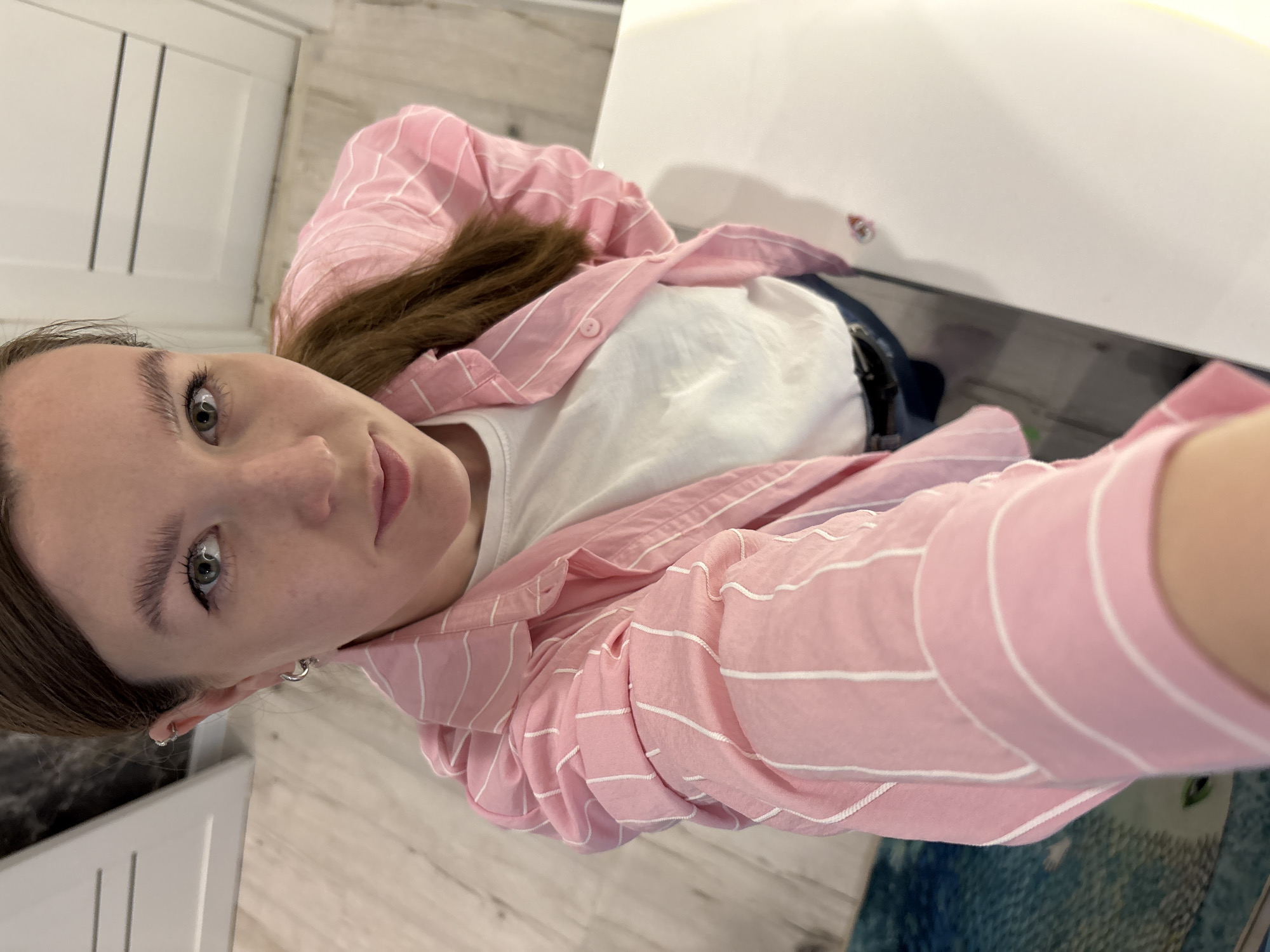 A high-angle selfie captures a young woman looking directly at the camera with a neutral expression. She has light-colored eyes, well-defined eyebrows, and brown hair. She is wearing a light pink, long-sleeved shirt with thin white horizontal stripes, unbuttoned over a plain white t-shirt. Multiple small silver hoop earrings are visible in her left ear. Dark blue lower garments, possibly jeans, are partially visible.
The setting appears to be an indoor space, likely a home in Izhevsk, Russia. The background features white cabinetry or furniture and a light-colored floor, possibly wood or tile. A dark surface or counter is visible behind her. A small, colorful sticker is faintly visible on a white surface in the upper right quadrant of the image. A patterned blue and green rug or mat is also visible in the lower right. The lighting is ambient and suggests daytime. There is no visible text in the image.