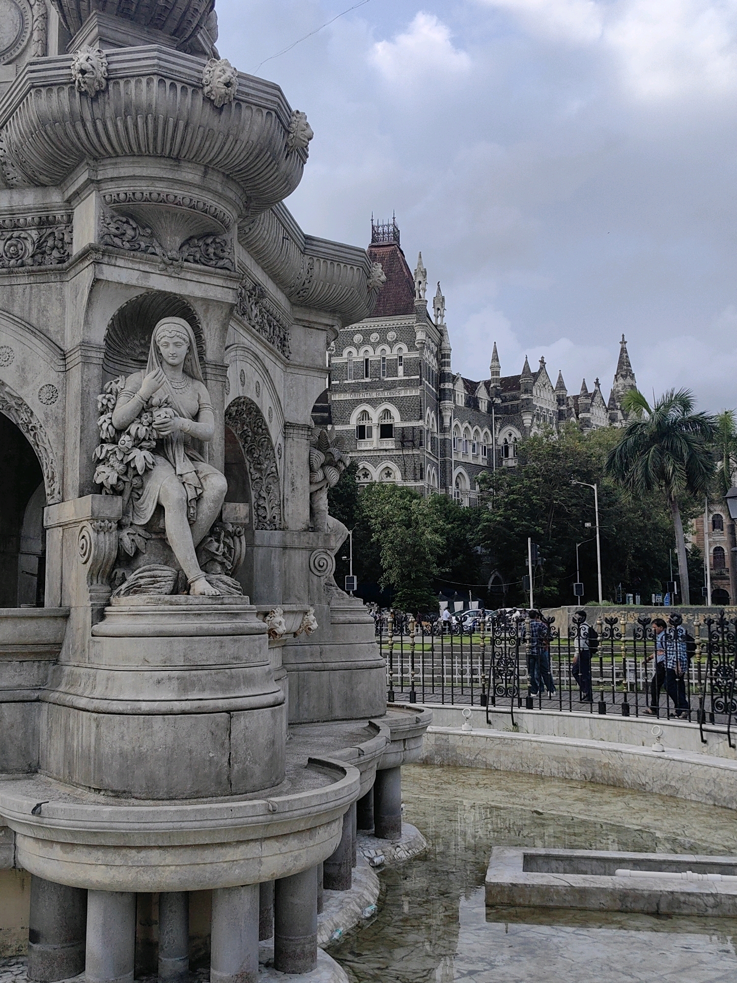 The image captures a view of the Flora Fountain in Mumbai, India. In the foreground, a section of the ornate stone fountain is visible, featuring a seated female figure adorned with a headdress and holding a bouquet of flowers. The fountain itself is made of intricately carved stone with lion heads and decorative elements. Behind the fountain, a large, Gothic-style building, likely the CSMT (Chhatrapati Shivaji Maharaj Terminus) or a similar heritage structure, rises prominently. Several people are walking on a pathway beyond a decorative black metal fence. The sky is overcast with clouds, and trees are interspersed between the fountain and the building, contributing to the urban landscape of Mumbai.