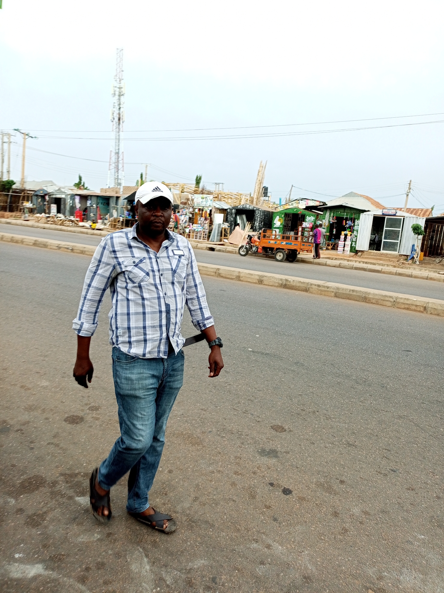 A man in a blue and white checkered shirt, blue jeans, and black sandals walks on a paved road. He wears a white baseball cap. A cell tower is visible in the background, along with several roadside stalls and buildings. A three-wheeled orange cart is parked near one of the stalls. The scene appears to be in a commercial area in Tempure, Nigeria, with a cloudy sky overhead.