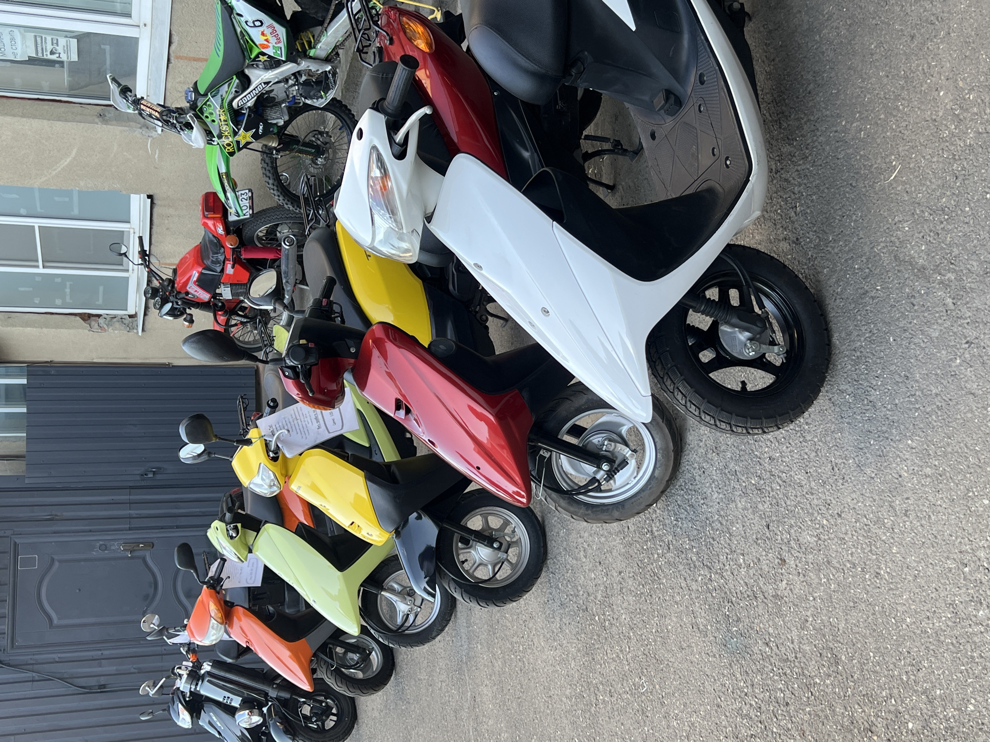 This media file captures a street scene in Krasnodar, Russia, featuring a collection of motorcycles and scooters parked in a row on an asphalt surface next to a building. The subjects include several two-wheeled vehicles arranged sequentially by color and type, suggesting a sales or rental business.
In the upper part of the image, a green and black dirt bike, adorned with "Red Bull," "Rockstar," and "ADDINOL" stickers, and the number "9," is visible. Behind it, a red street motorcycle with a partially visible Russian license plate "КО 23" is parked. The main focus then shifts to a line of scooters: a white scooter is prominent on the right, followed by a yellow, red, light green, orange, and black scooter, and more further down the line. Several scooters have white informational papers, likely price lists or descriptions, attached to their handlebars.
The setting includes building facades on the left side of the frame. One building is light-colored with windows, one of which displays some difficult-to-read Cyrillic text. Further down, a large, dark, paneled door is visible. The ground is paved with asphalt. The bright lighting indicates that the photograph was taken during the day under clear weather conditions. No people are visible in the scene.