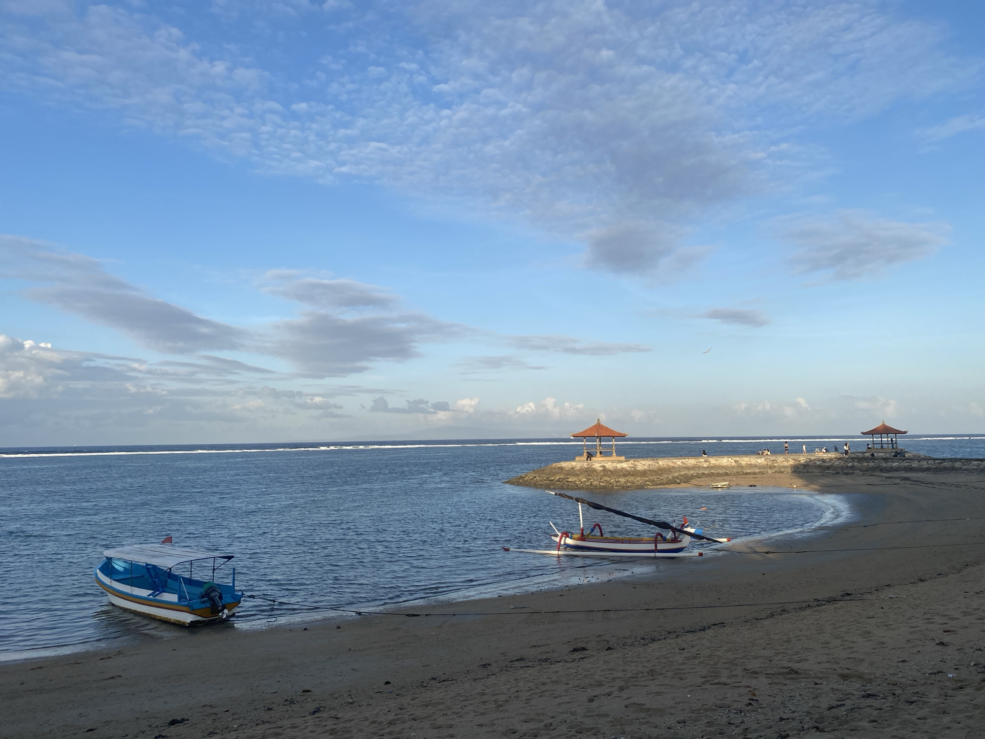 This is a full shot of a beach scene in Denpasar, Indonesia, taken at eye-level. The sky is blue with scattered clouds, and the sun is low in the sky, casting a gentle light on the scene. In the foreground, there is a sandy beach with two boats moored near the shore. The boat on the left is a small, white and blue motorboat with a canopy. The boat on the right is a traditional Balinese fishing boat with outriggers and a red and white striped hull. In the middle ground, there is a stone jetty with two gazebos. Several people are visible on the jetty, some standing and some sitting. The water is a calm, blue sea with a white line of surf in the distance. The overall mood of the image is peaceful and serene.
