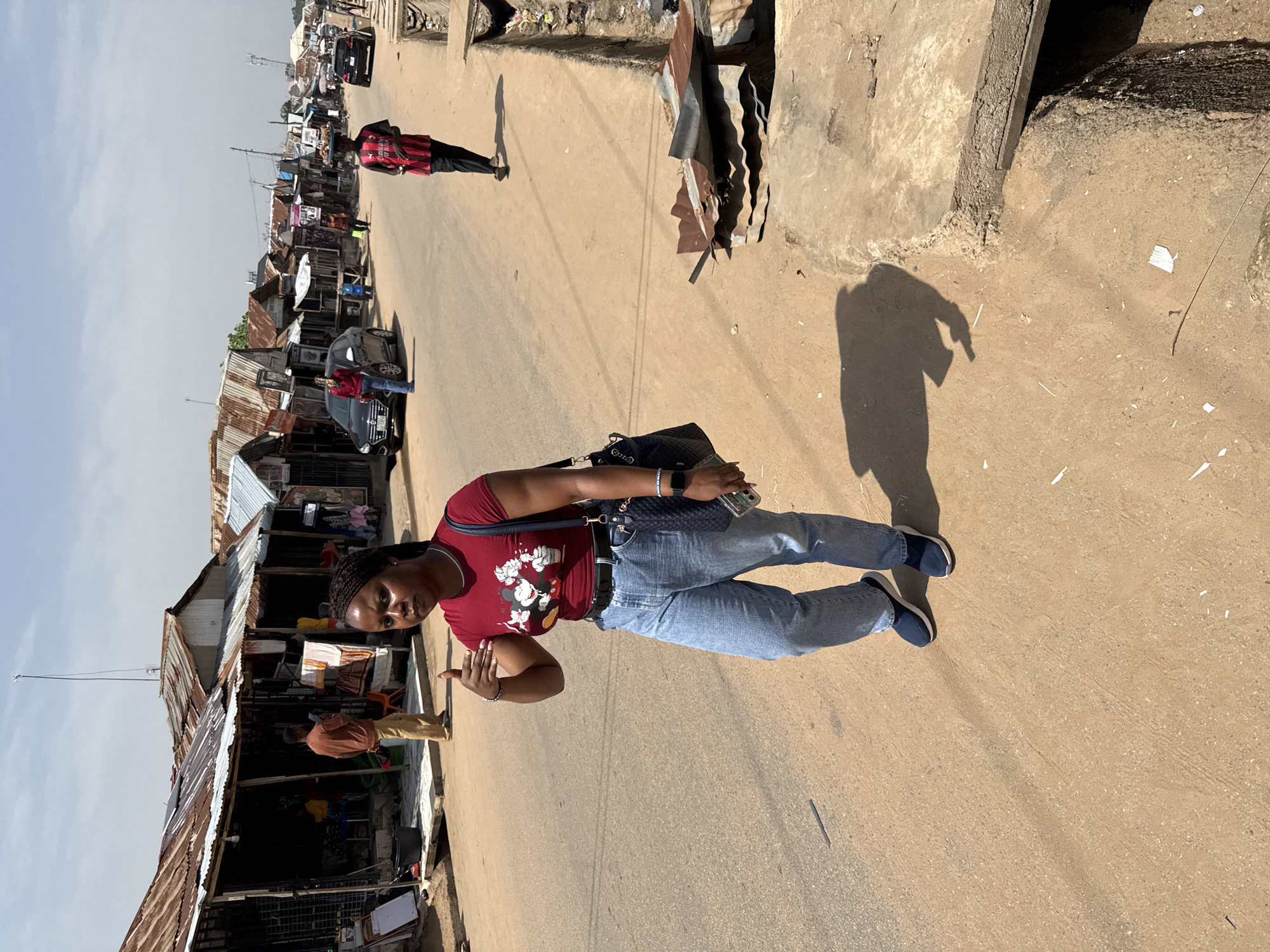 A woman with braided hair walks down a dusty street in Abuja, Nigeria. She is wearing a red t-shirt with a cartoon print, blue jeans, and blue sneakers. A dark blue purse hangs from her shoulder. She is gesturing with her left hand, and her right hand holds a smartphone. Behind her, several market stalls with corrugated metal roofs line the street. A man in a red and black striped shirt walks in the background carrying a tray of goods on his head. Cars and other pedestrians are visible further down the street. The ground is sandy and uneven, with visible debris and drainage ditches. The sky is clear and blue.