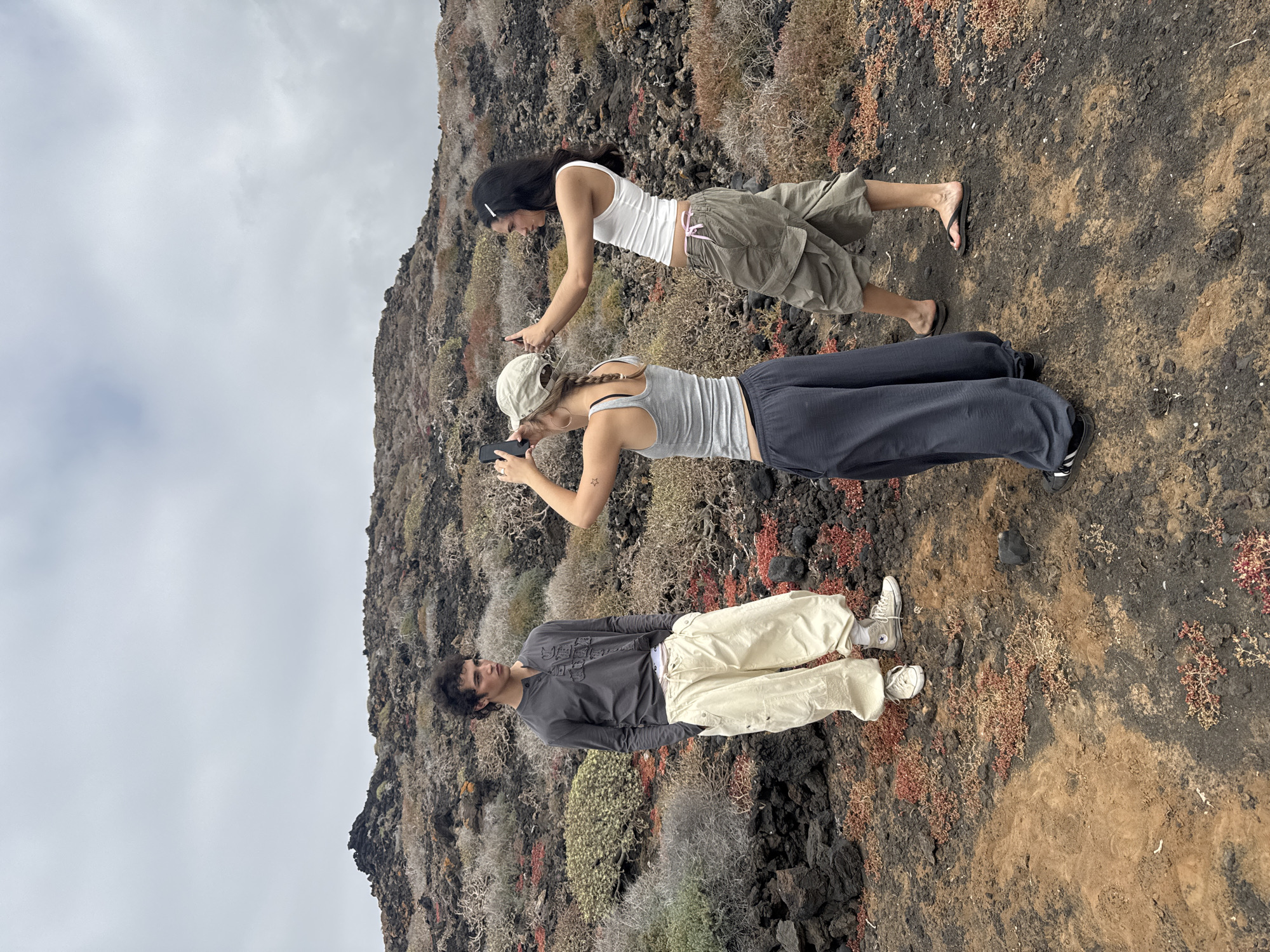 In Tinajo, Spain, three individuals stand on a rocky, volcanic landscape with sparse vegetation under a cloudy sky. One person, with long dark hair, is wearing a white tank top and olive-colored shorts, appearing to be using a mobile phone. Another person, with braided hair and wearing a baseball cap, is also using a phone. The third individual wears a long-sleeved dark top and light-colored trousers. The surroundings consist of dark rocks and patches of brown soil.