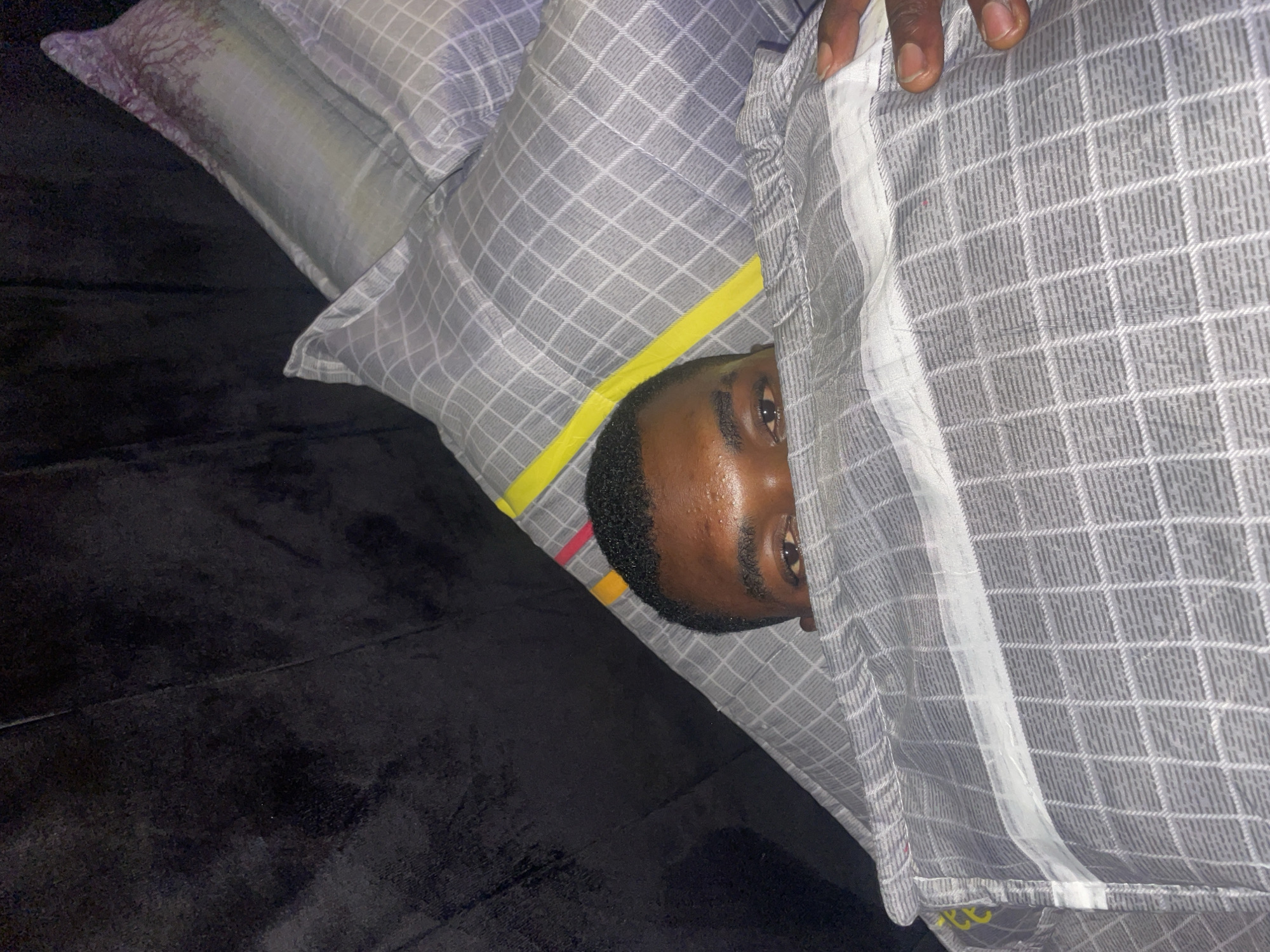 A person with dark skin and short dark hair is partially visible, peeking out from under a grey and white checkered blanket. The person's forehead is shiny, suggesting perspiration. The person is lying on a black surface, likely a bed or couch. Two pillows, one with a light grey checkered pattern and a yellow accent, and another with a tree design, are visible near the person's head. A hand is partially seen holding the blanket near the top right corner of the image. The location of the photo is unknown, but I am able to say it is Jos, Nigeria.