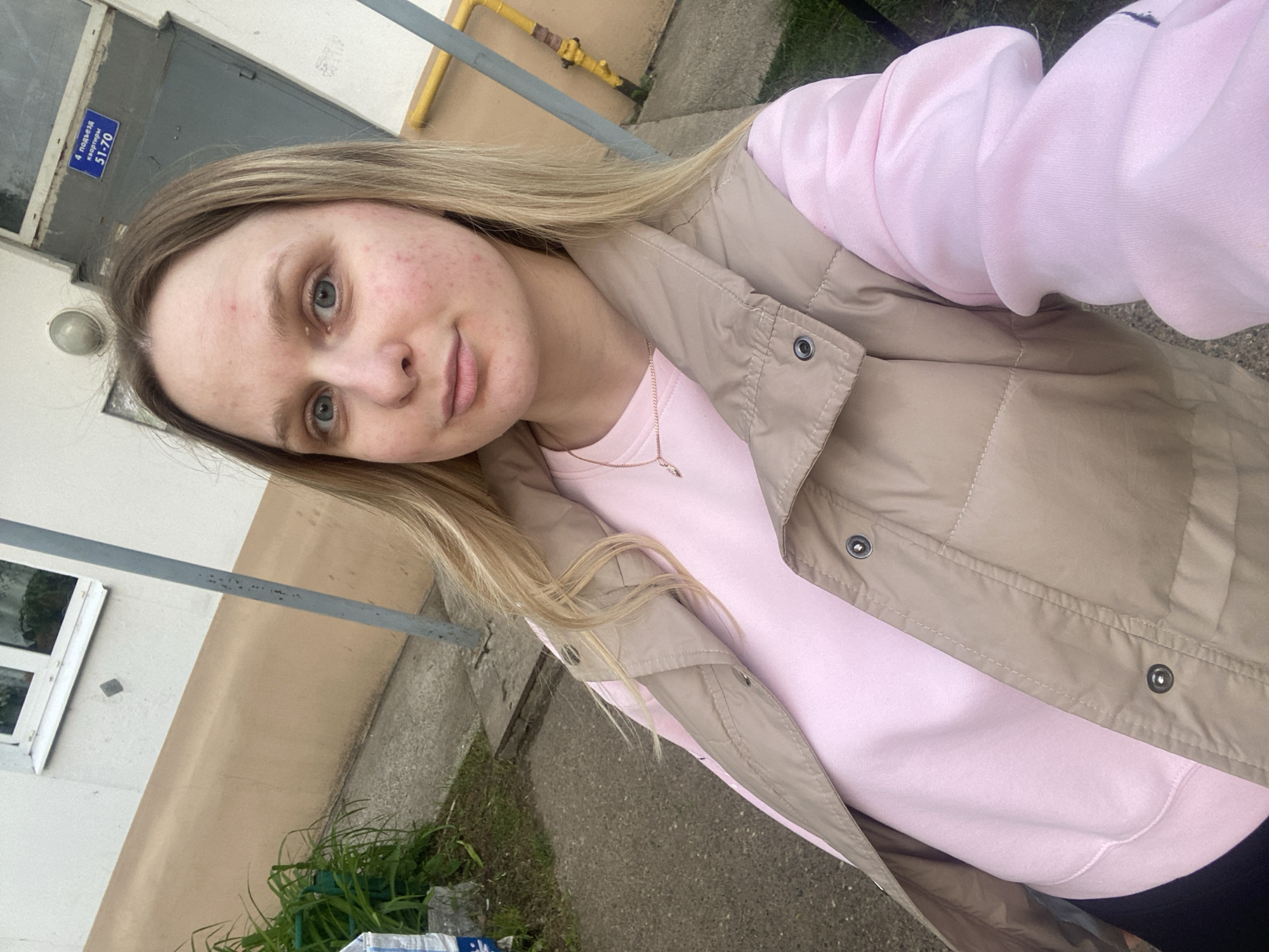 A light-skinned woman, appearing to be in her 20s or early 30s, is centered in a daytime, eye-level selfie, looking directly into the camera with a neutral expression. Her blonde hair is shoulder-length, slightly parted, and frames her face. She has blue eyes, and her skin shows visible redness and blemishes across her forehead, cheeks, and chin. She is wearing a light pink crew-neck long-sleeve sweatshirt beneath a beige or tan quilted puffer vest with snap buttons visible down the front. A thin gold-colored chain necklace with a small, indistinct pendant is visible around her neck. The bottom left of the frame shows a sliver of black fabric, likely part of her lower attire.
The setting is outdoors in front of an older residential building in Unknown, Unknown. The building facade behind her is two-toned: white or off-white on the upper section and a lighter tan or light brown on the lower half. A white-framed window is visible on the upper left. To the woman's upper right, a dark grey metal door, likely an entrance to a residential building, is visible. Above this door, a dome-shaped grey light fixture and a rectangular blue sign with white Cyrillic text are present. The sign reads "4 подъезд" (4th entrance) and "квартиры 51-70" (apartments 51-70). A yellow gas pipe is visible running along the tan section of the wall to the right of the woman. A grey metal pole extends vertically from the lower right to the mid-upper right of the image. The ground consists of a grey concrete sidewalk or pathway, with green grass and leafy plants along the left edge. A partially obscured white object, possibly a bag, sits on the ground near the plants.